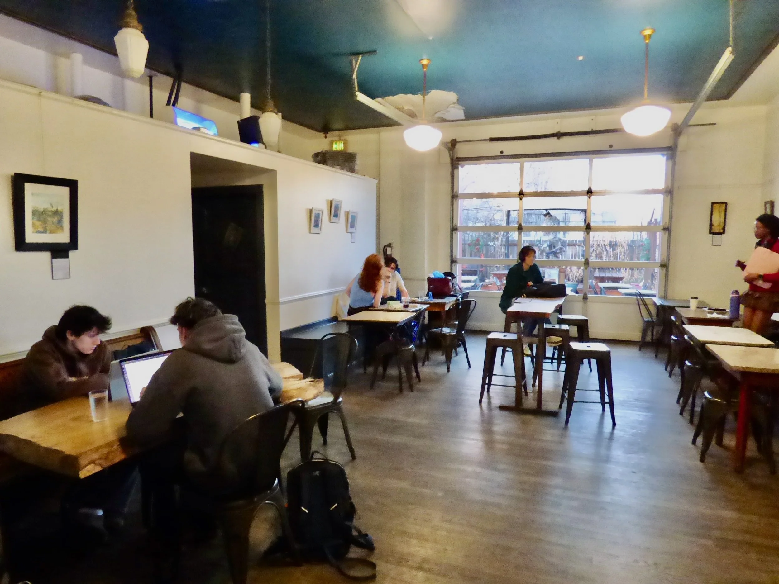 A cozy cafe interior with wooden floors, white walls decorated with small framed pictures, and large windows providing natural light. There are several tables, with a few people working or chatting  at St. Mark's Coffeehouse and Bakery, Denver, CO.