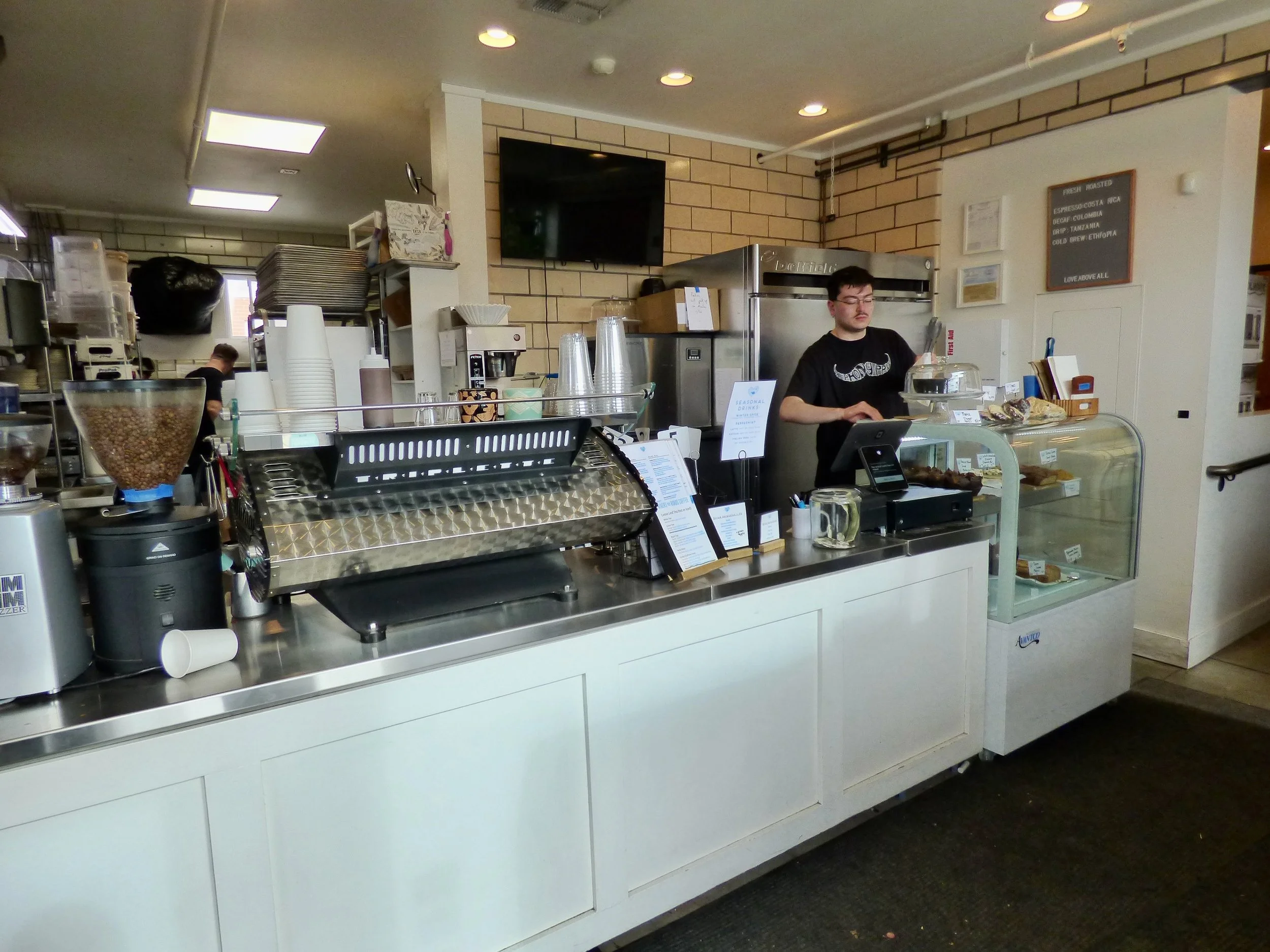 Inside of a coffee shop with a barista behind the counter preparing drinks, coffee equipment, a glass display case with baked goods, and a menu board on the wall.