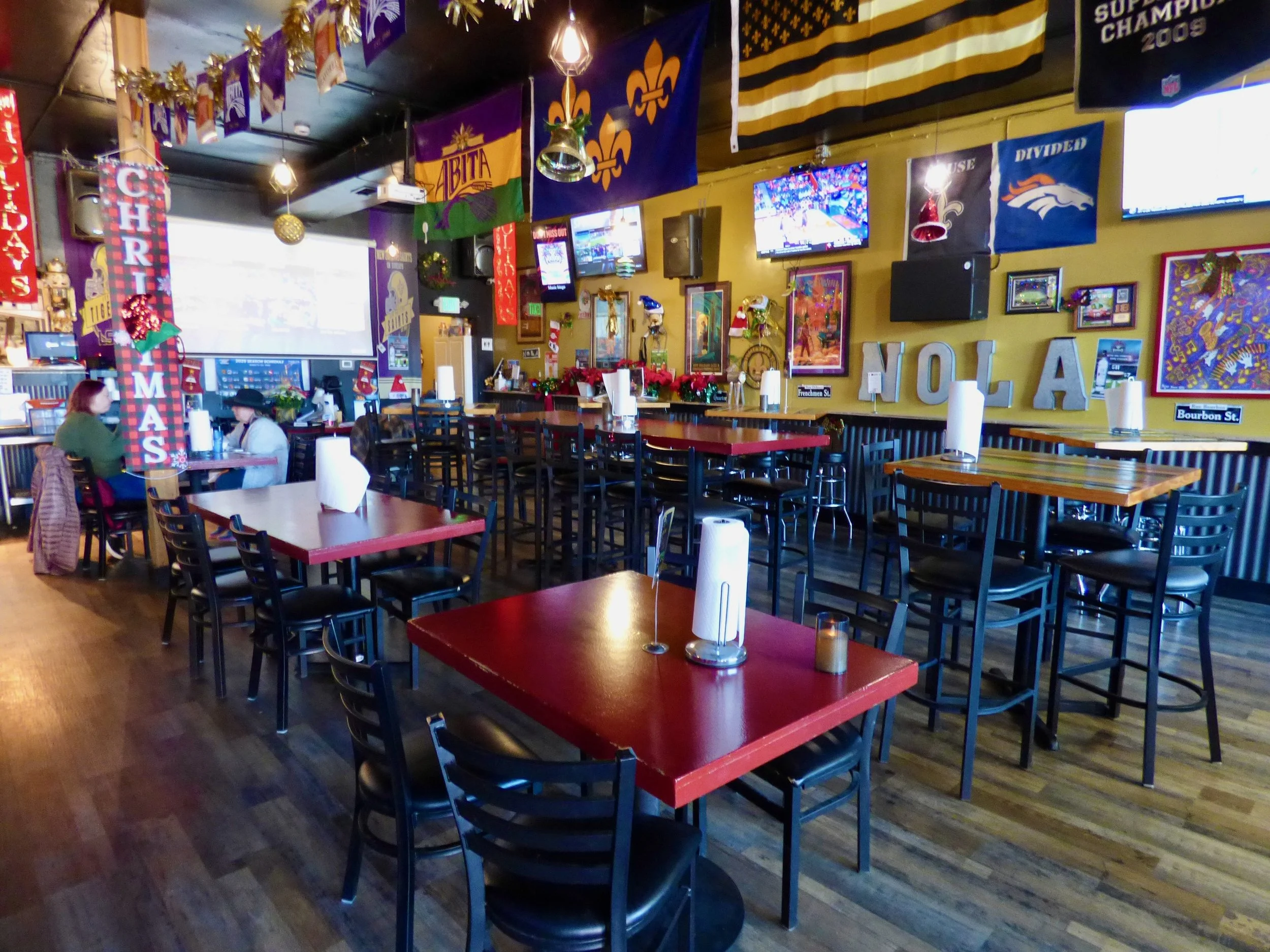 A sports bar decorated for Christmas with tables, chairs, and wall decorations including flags, banners, and televisions at NOLA Voodoo Tavern, Denver, CO 80205.