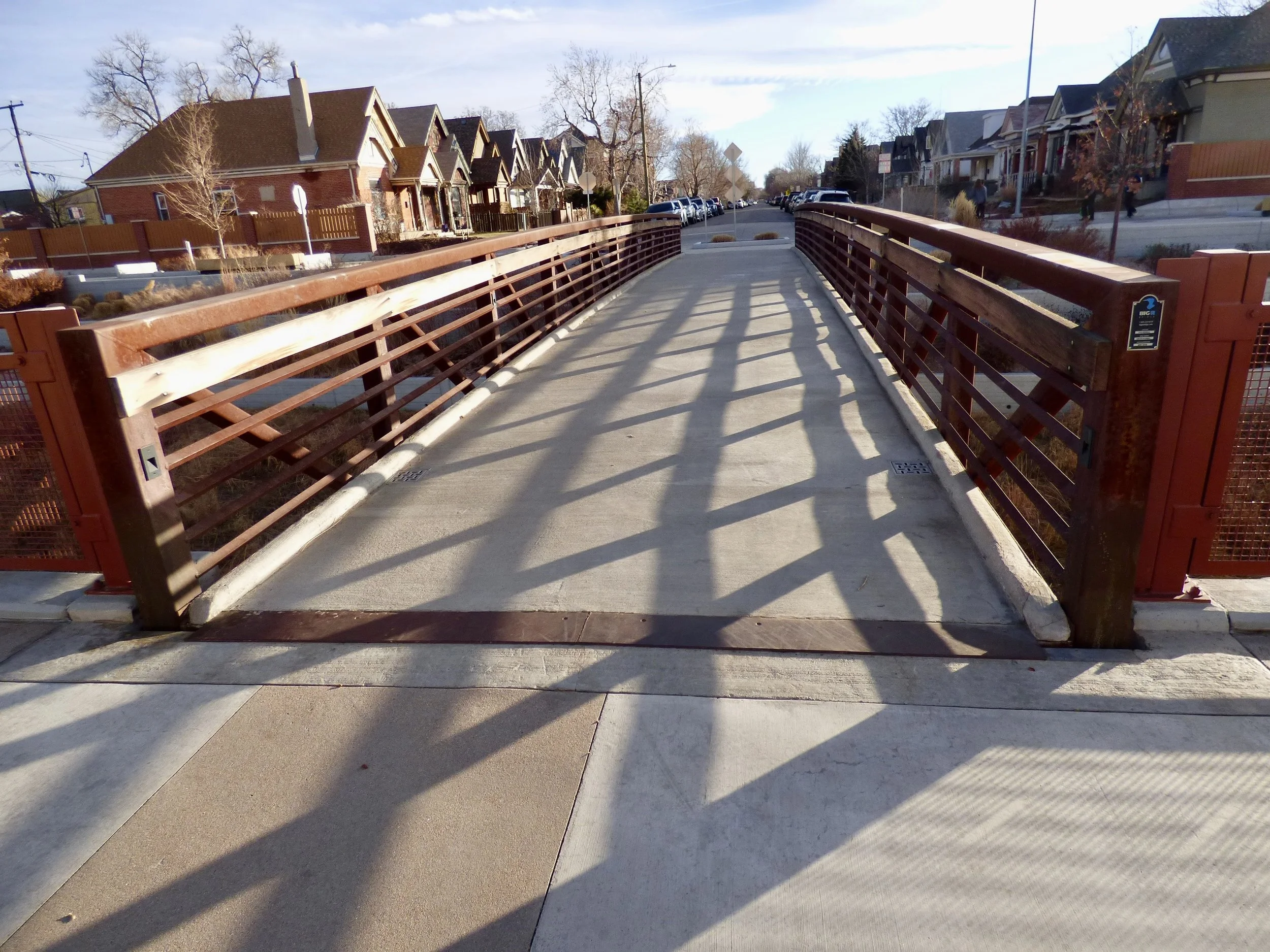 A pedestrian bridge with wooden and metal railings over a small waterway, with residential houses in the background and shadows cast on the walkway in 39th Ave Greenway, Denver, CO 80205.