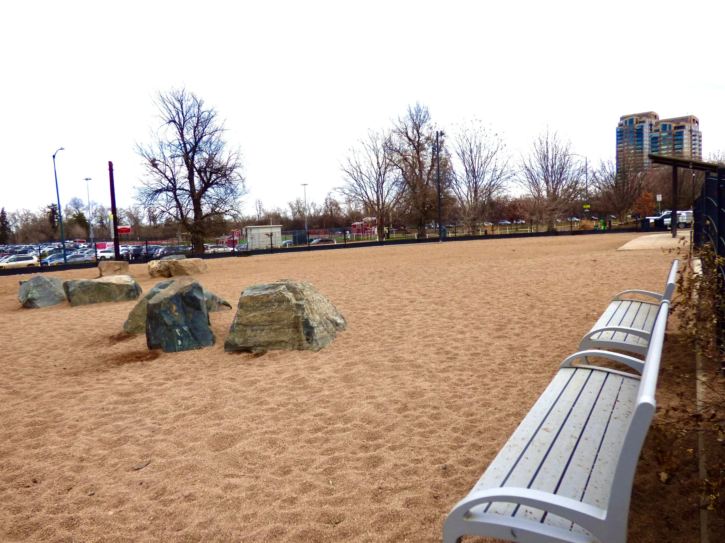 Empty sandy park with large rocks, trees without leaves, benches, and a parking lot in the background, with buildings and cloudy sky.