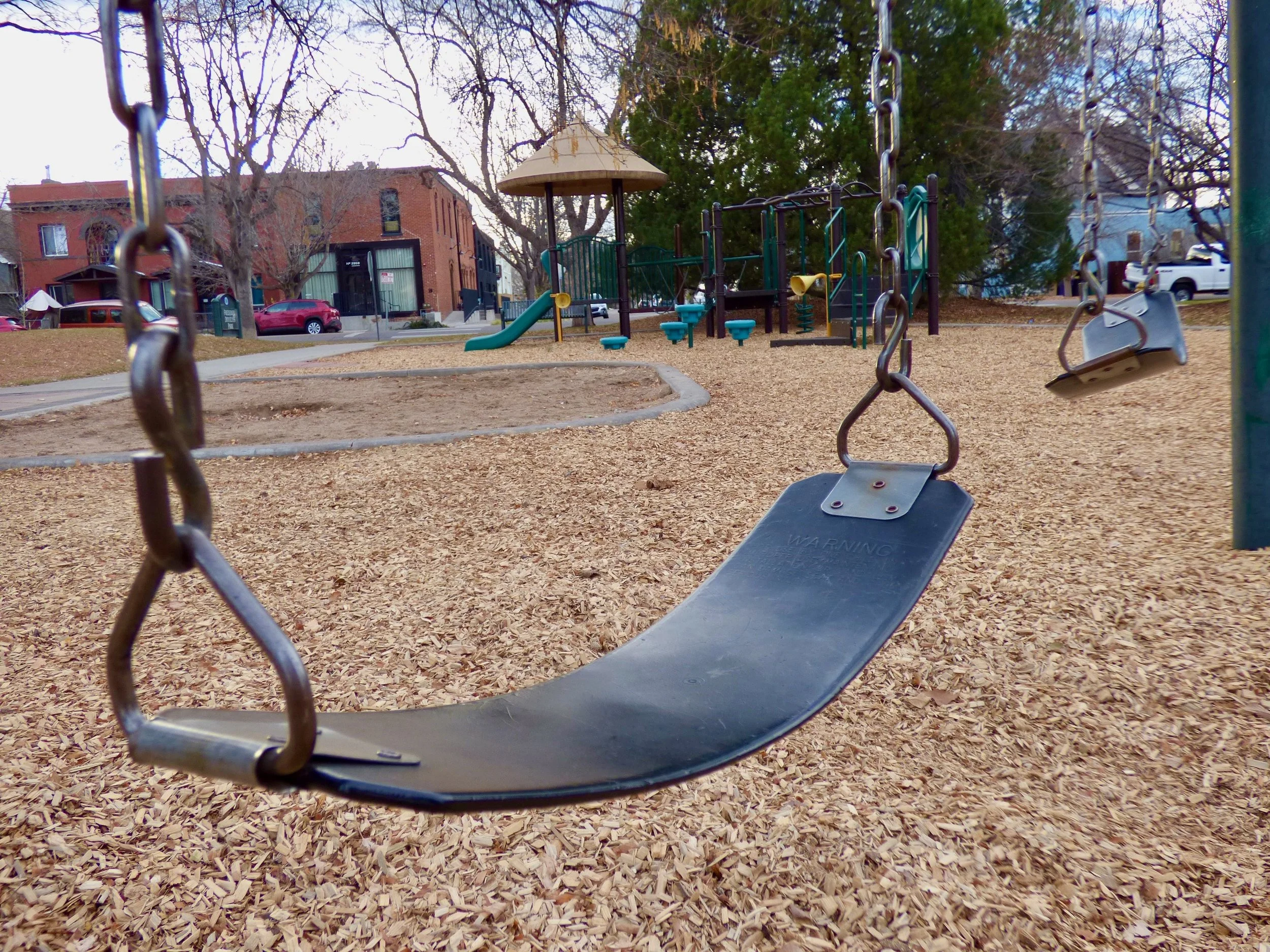 Empty playground swing set with wood chip ground cover, and a play structure with a slide and climbing bars in the background, with trees and houses visible at Frederick Douglas Park, Denver, CO 80205.