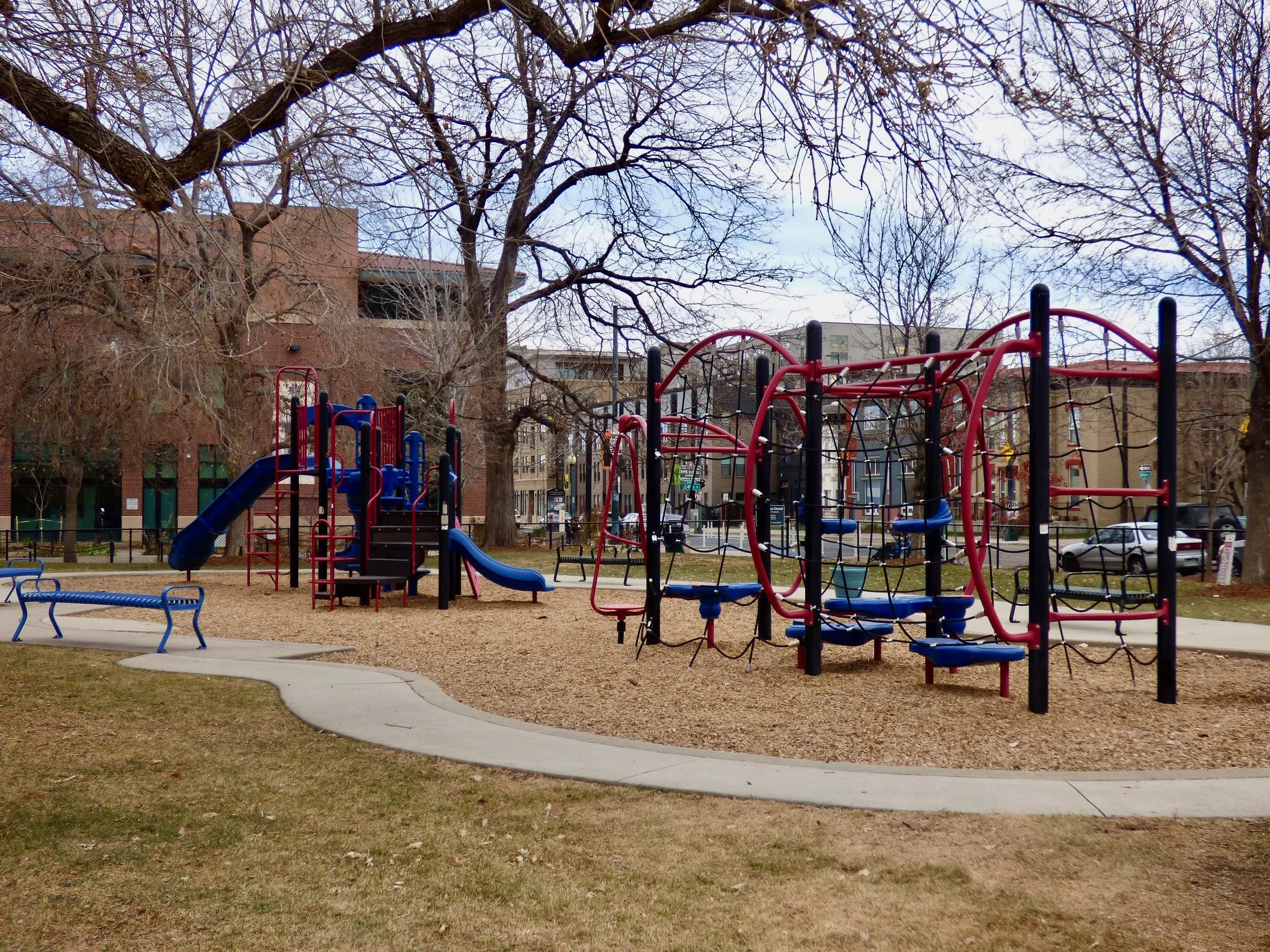 A park with playground equipment, including slides and climbing structures, surrounded by trees and a grassy area, with buildings in the background in Sonny Lawson Park, Denver, CO 80205.