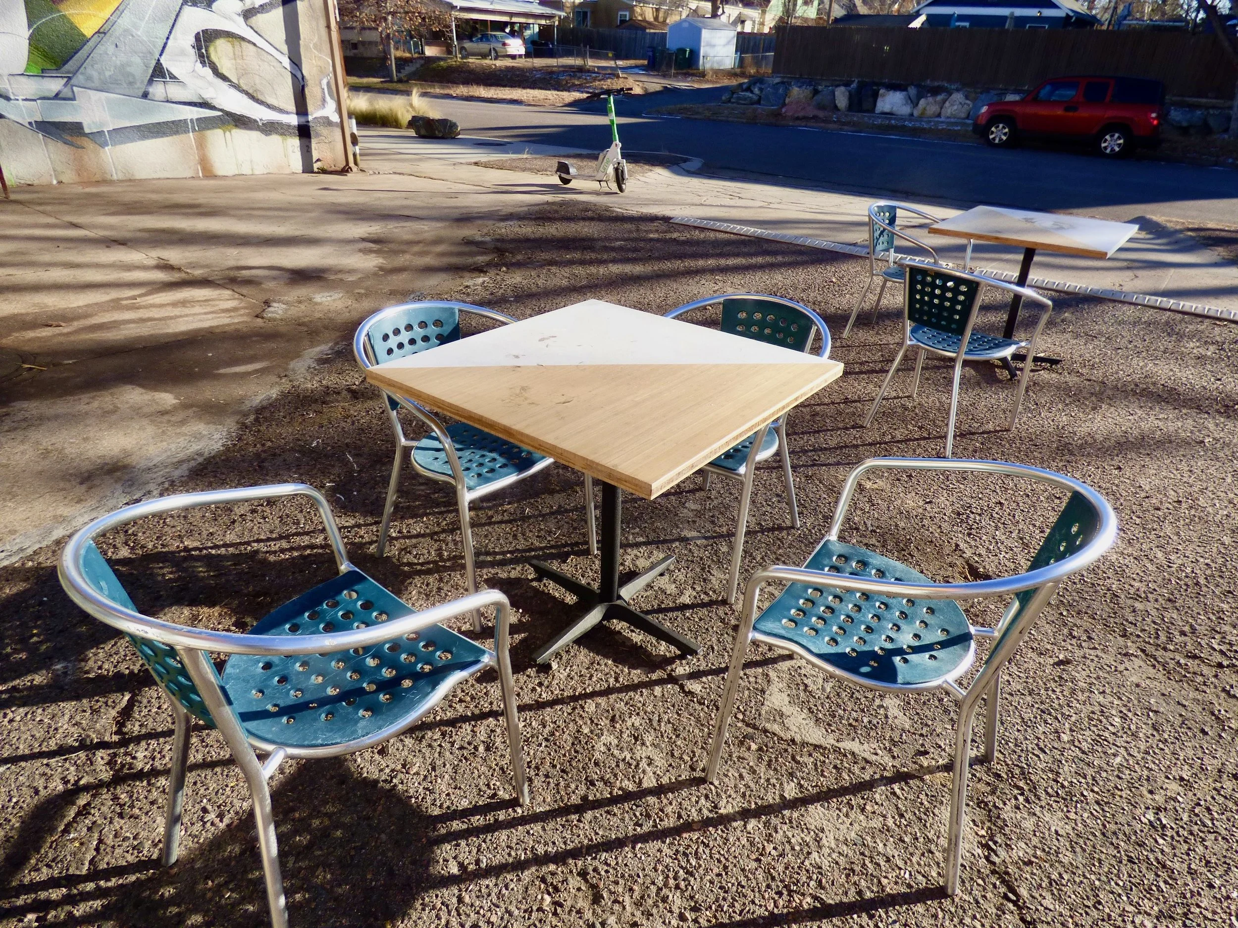 Outdoor seating area with four blue chairs and two tables with wooden tops, situated on a gravel surface. In the background, there is a street with a parked red vehicle, a graffiti-covered wall, and a scooter.