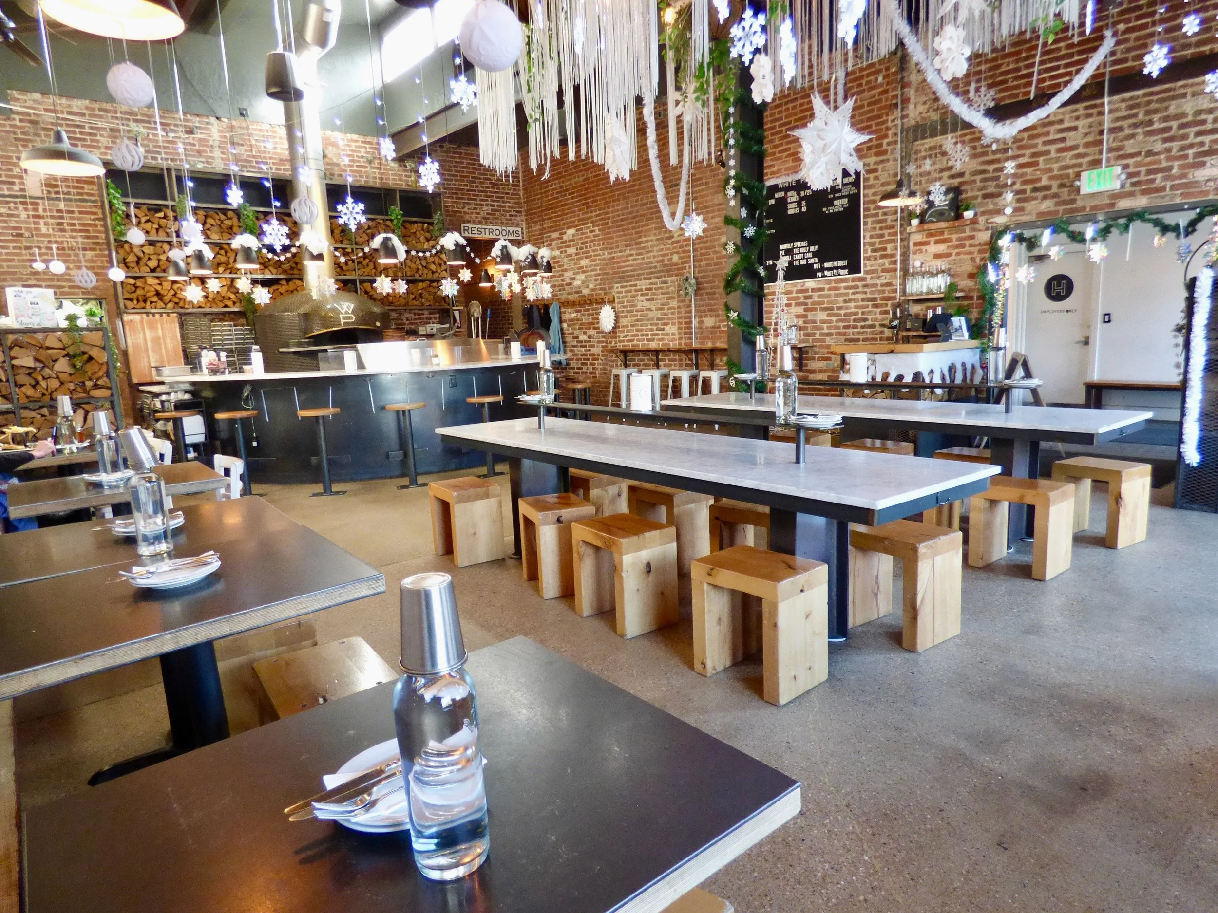 Decorated restaurant interior with hanging paper snowflakes and white garlands, brick walls, wooden and black tables with water glasses and silverware, and a bar area in the background.