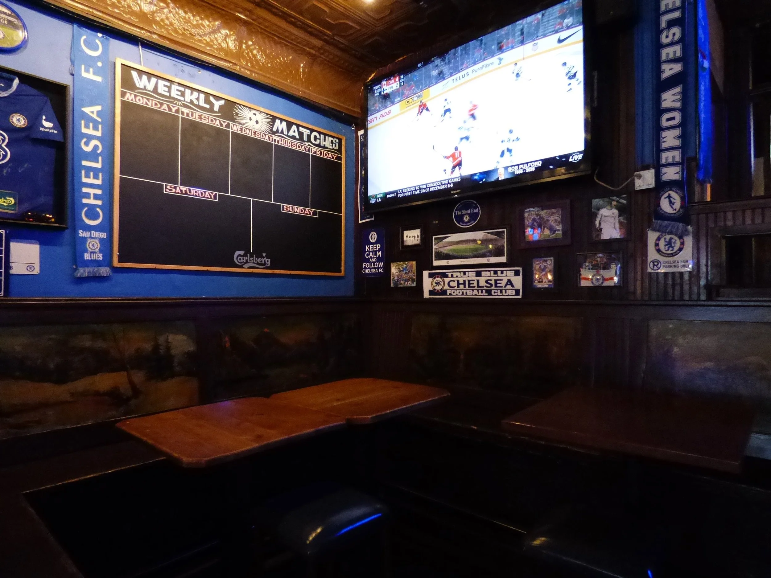 Interior of a sports bar with a large flat-screen TV showing an ice hockey game, surrounded by Chelsea football club memorabilia and a weekly sports schedule board on dark wooden walls at The British Bulldog, Denver, CO 80205.