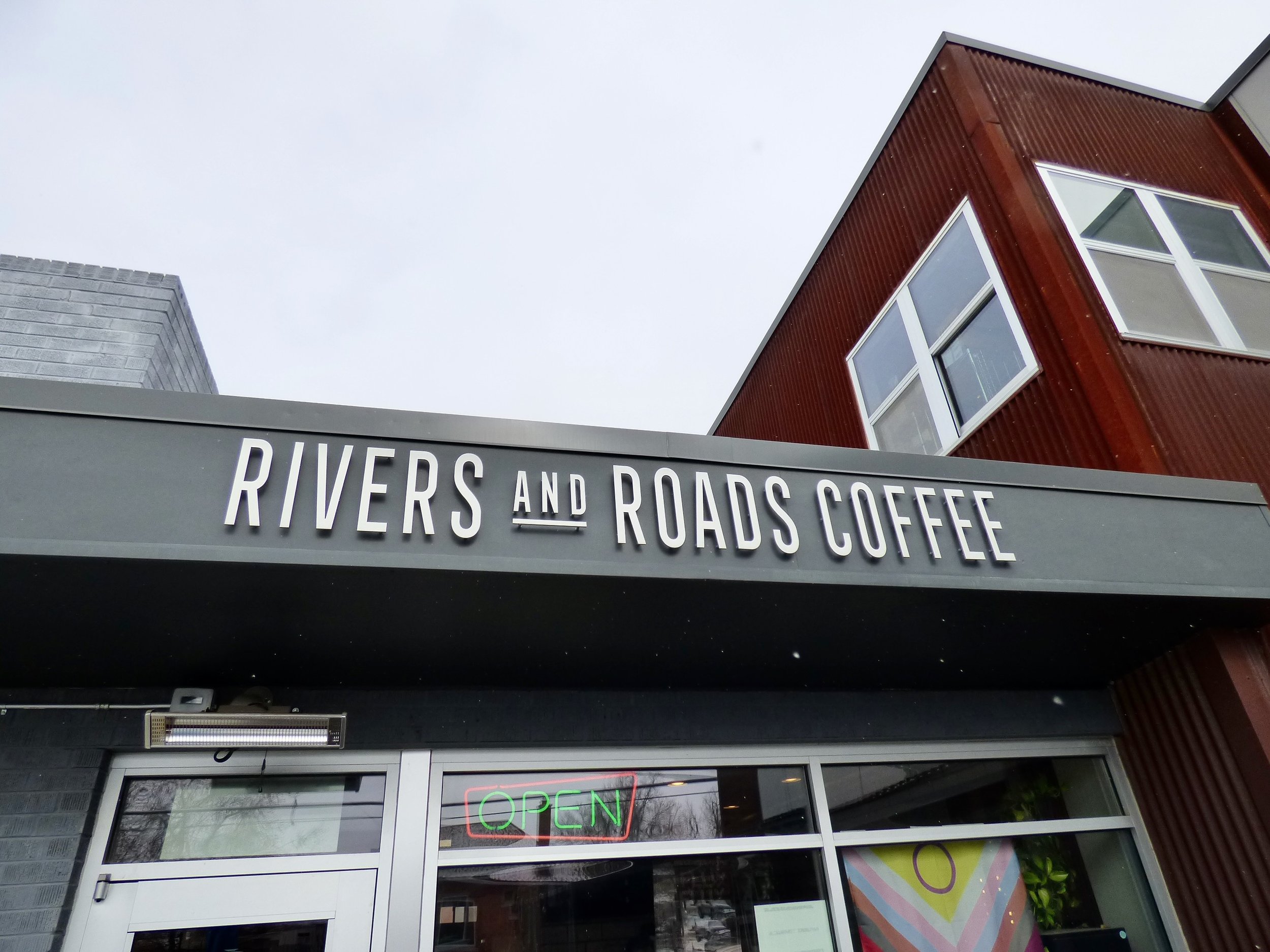 Exterior view of RIVERS AND ROADS COFFEE shop with an open sign in the window and a cloudy sky above.