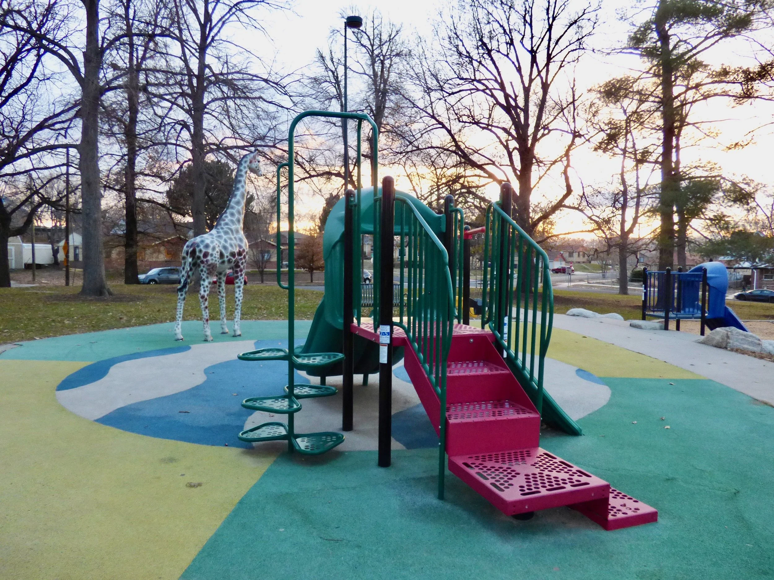 Colorful playground with a plastic slide, stairs, and a climbing area, situated on a surface with painted shapes and a plastic giraffe statue nearby, set in a park with leafless trees at sunset.