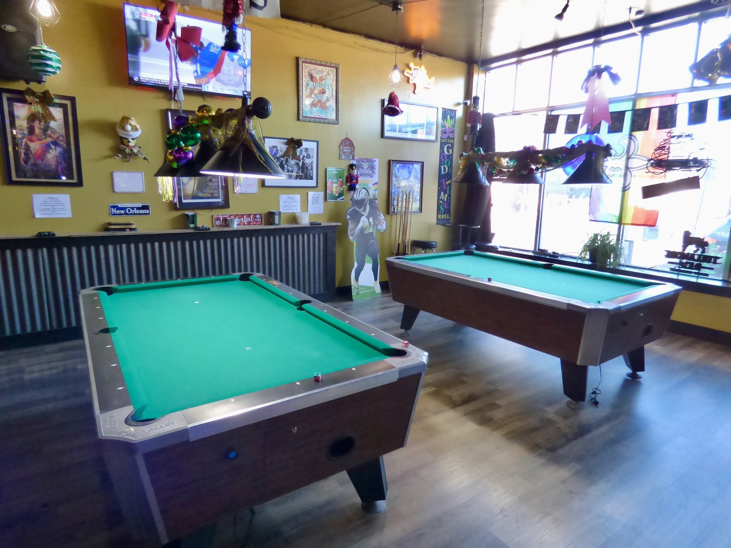 Two pool tables in a decorated bar or game room with yellow walls, wall art, a TV, and large windows letting in natural light at NOLA Voodoo Tavern, Denver, CO 80205.