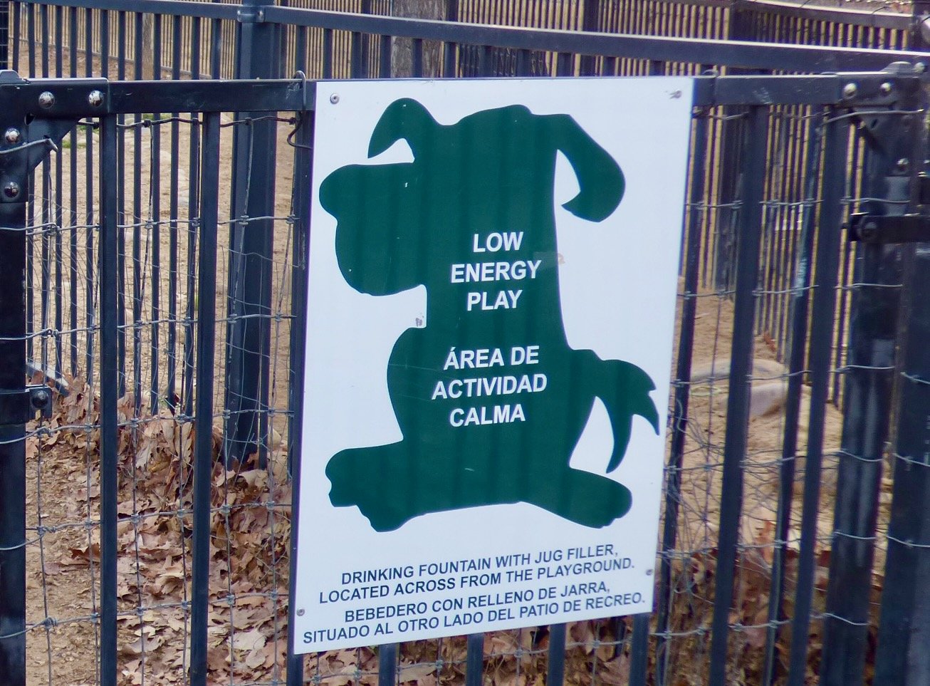 Sign on a fence indicating it is a low energy play area with a dog shape and bilingual text about a drinking fountain.