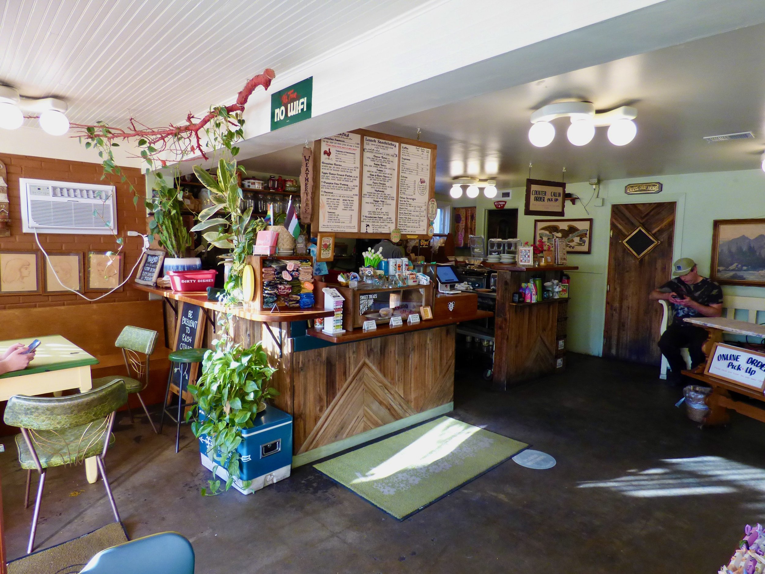 Interior of a cozy cafe with a wooden counter, menu boards, paintings, plants, and a customer seated near the door at Weathervane Cafe, Denver, CO.