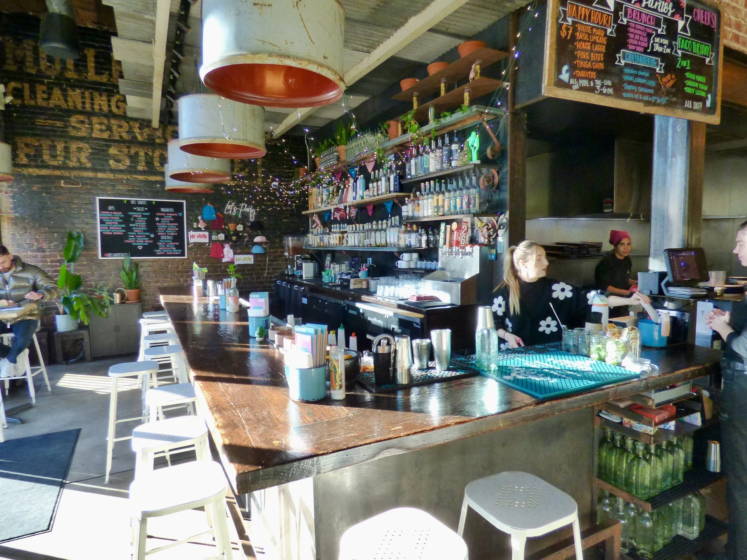 Inside a cozy cafe with a wooden bar counter, baristas preparing drinks, colorful signage, string lights, and seating area with white chairs and potted plants at Dos Santos, Denver, CO.