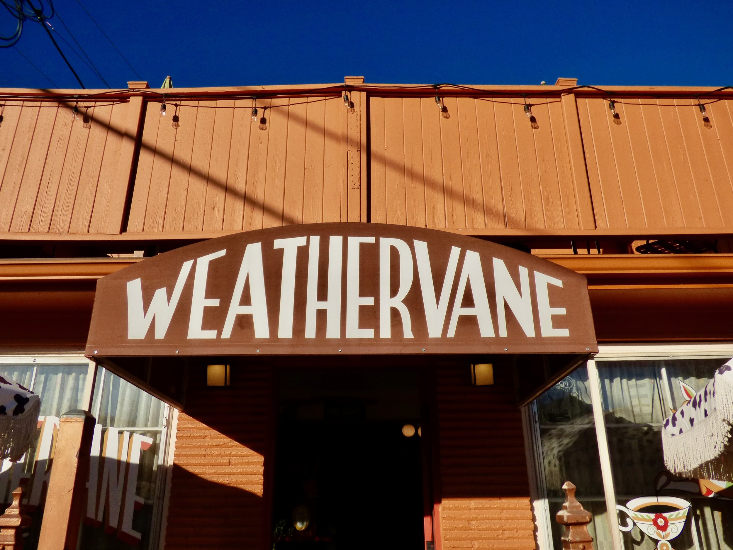 The image shows the exterior of a building with a large sign reading 'WEATHERVANE' in capital letters, with a bright blue sky overhead at Weathervane Cafe, Denver, CO.