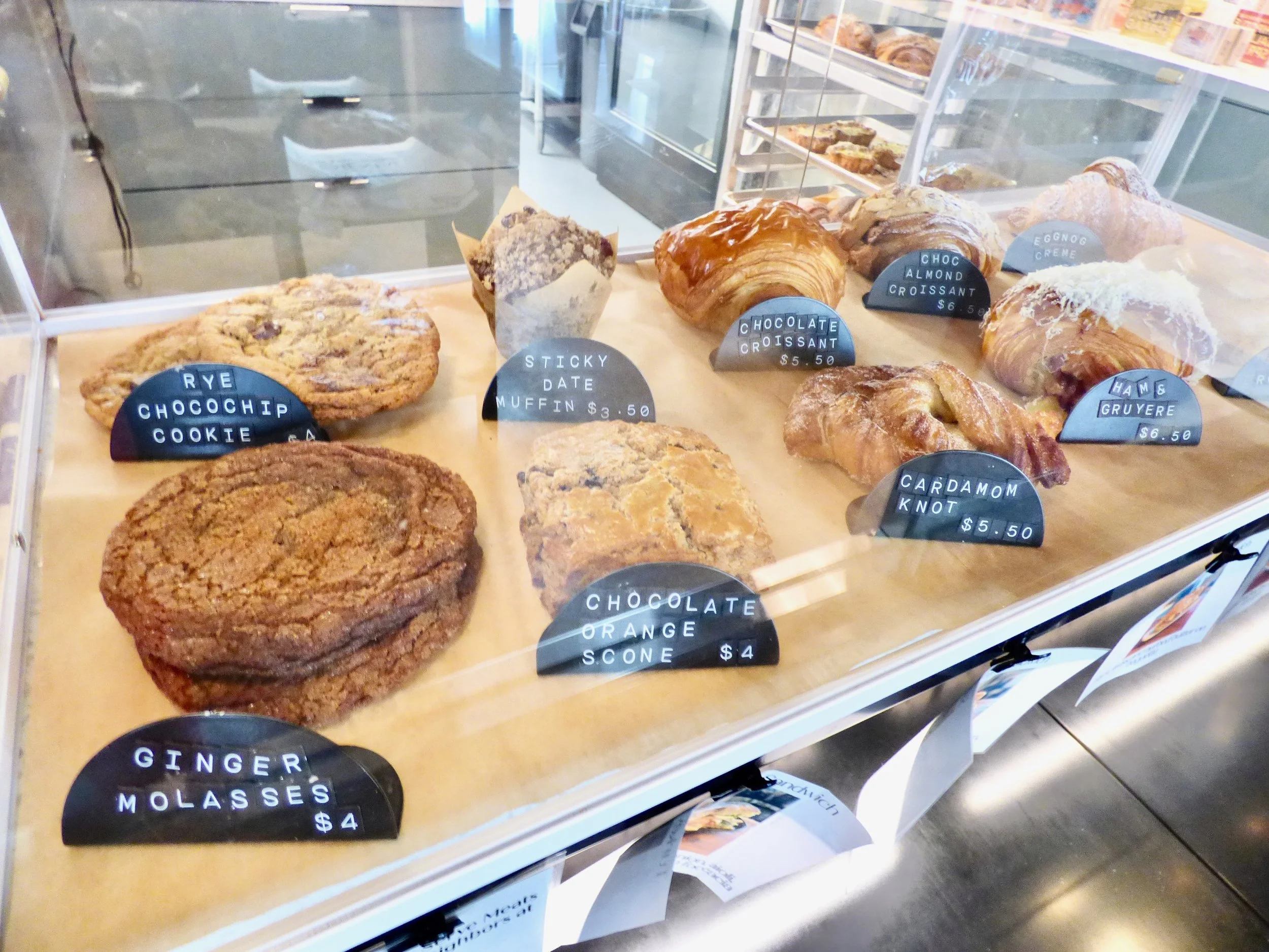 Assorted baked goods displayed in a bakery case, including cookies, muffins, croissants, and scones with labels and prices at Elemental Bakery & Coffeehouse, Denver, CO 80205.
