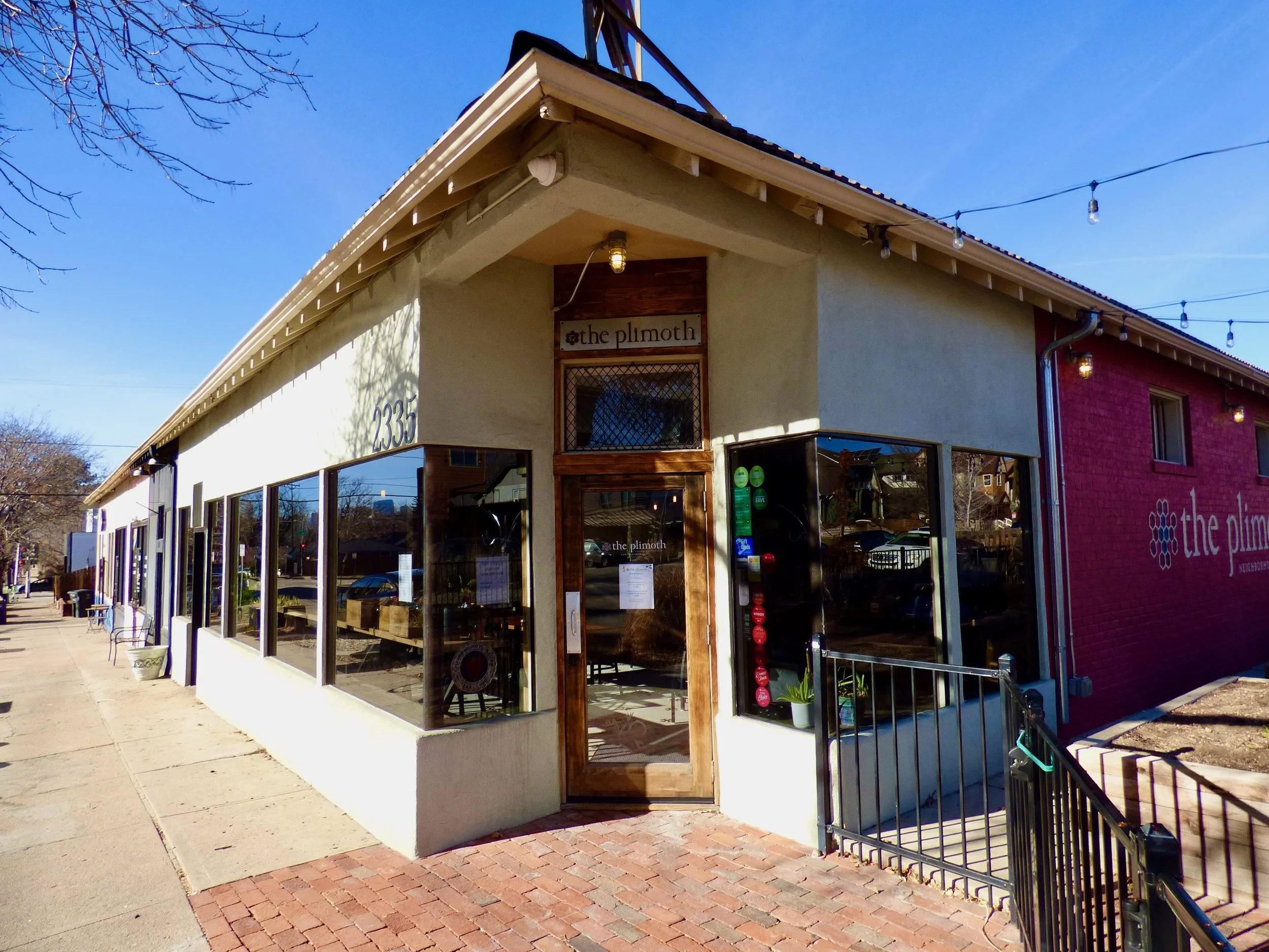 A corner view of a small café called 'the plmoth' with large front windows, a wooden door, a sign above the door, and string lights overhead at The Plimoth, Denver, CO 80205.