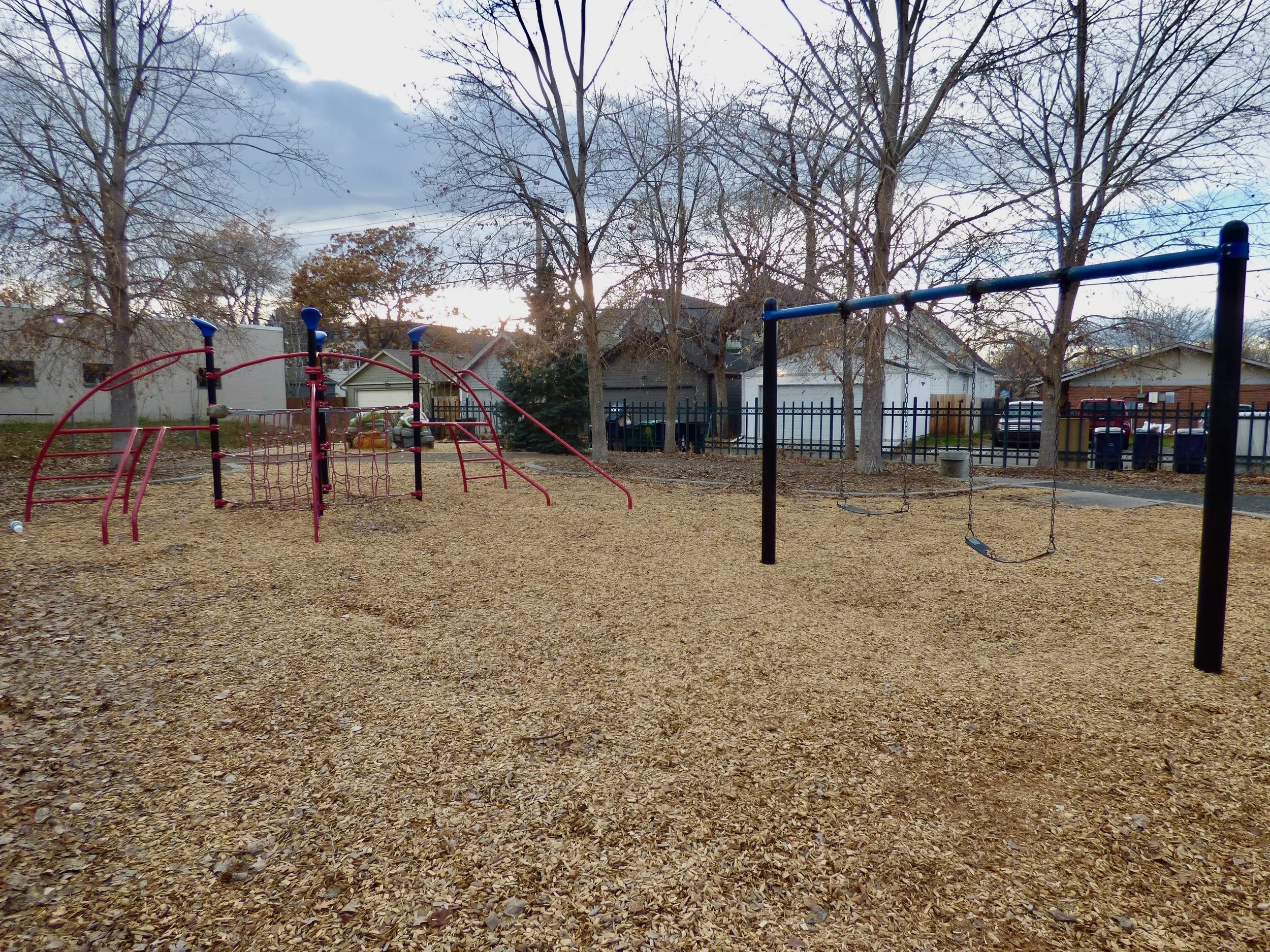 Empty playground with a climbing structure and swings, surrounded by trees and houses under a cloudy sky at Madame C.J. Walker Park, Denver, CO 80205.