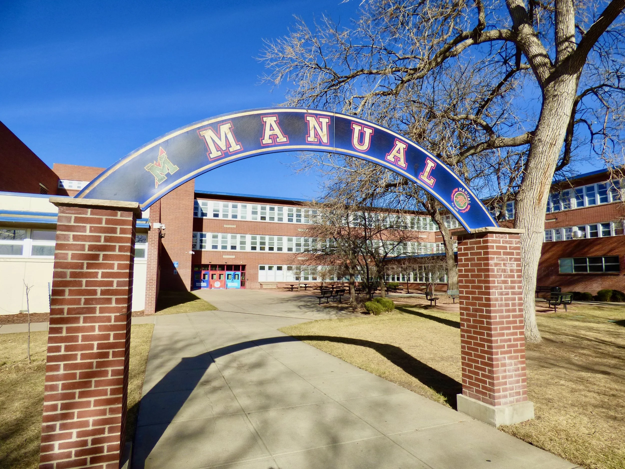 School entrance with an archway sign reading 'MANUAL' in front of a brick building, leafless trees, and benches on a sunny day at Manual High School, Denver, CO 80205.