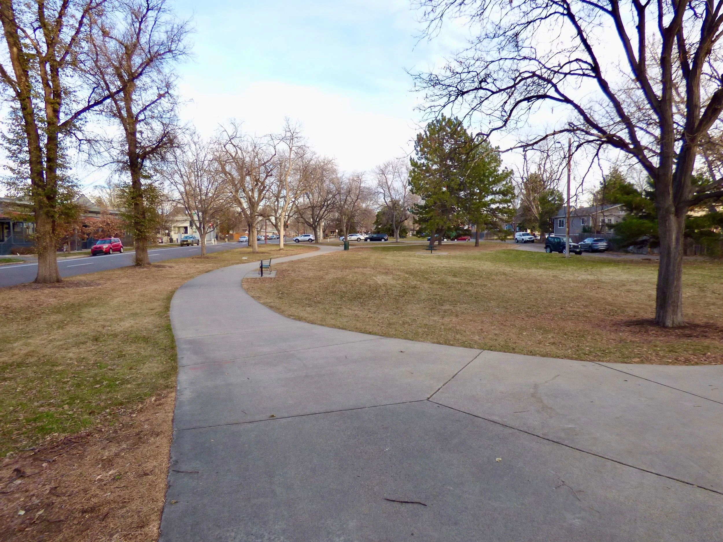 A curved concrete sidewalk running through a park with leafless trees, benches, and a grassy area, with houses and cars in the background under a clear sky at George Morrison Sr. Park, Denver, CO 80205.