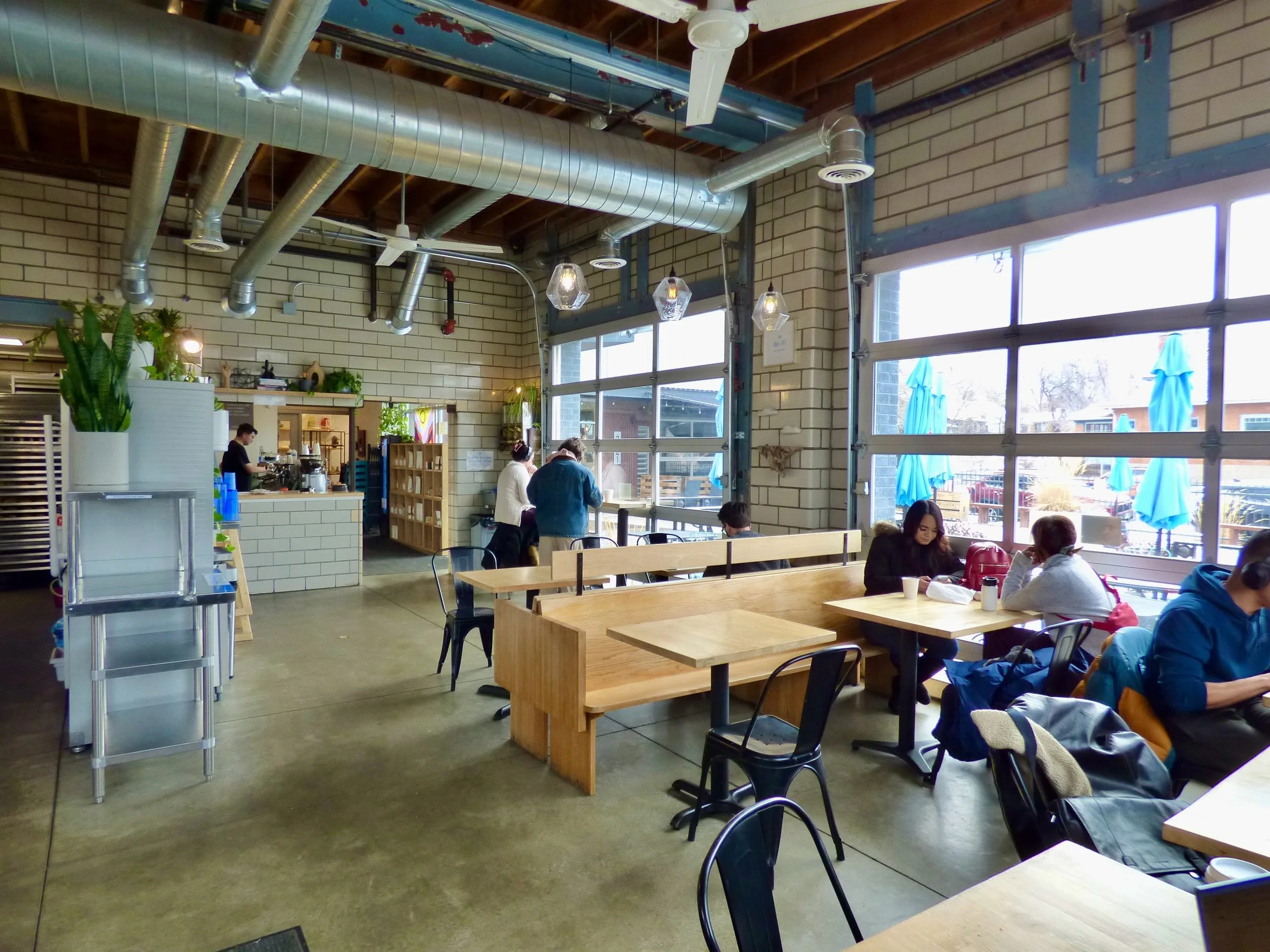 Interior of a modern cafe with wooden tables, black chairs, and large windows; a few customers sit and work or chat, with some staff behind the counter near the entrance.