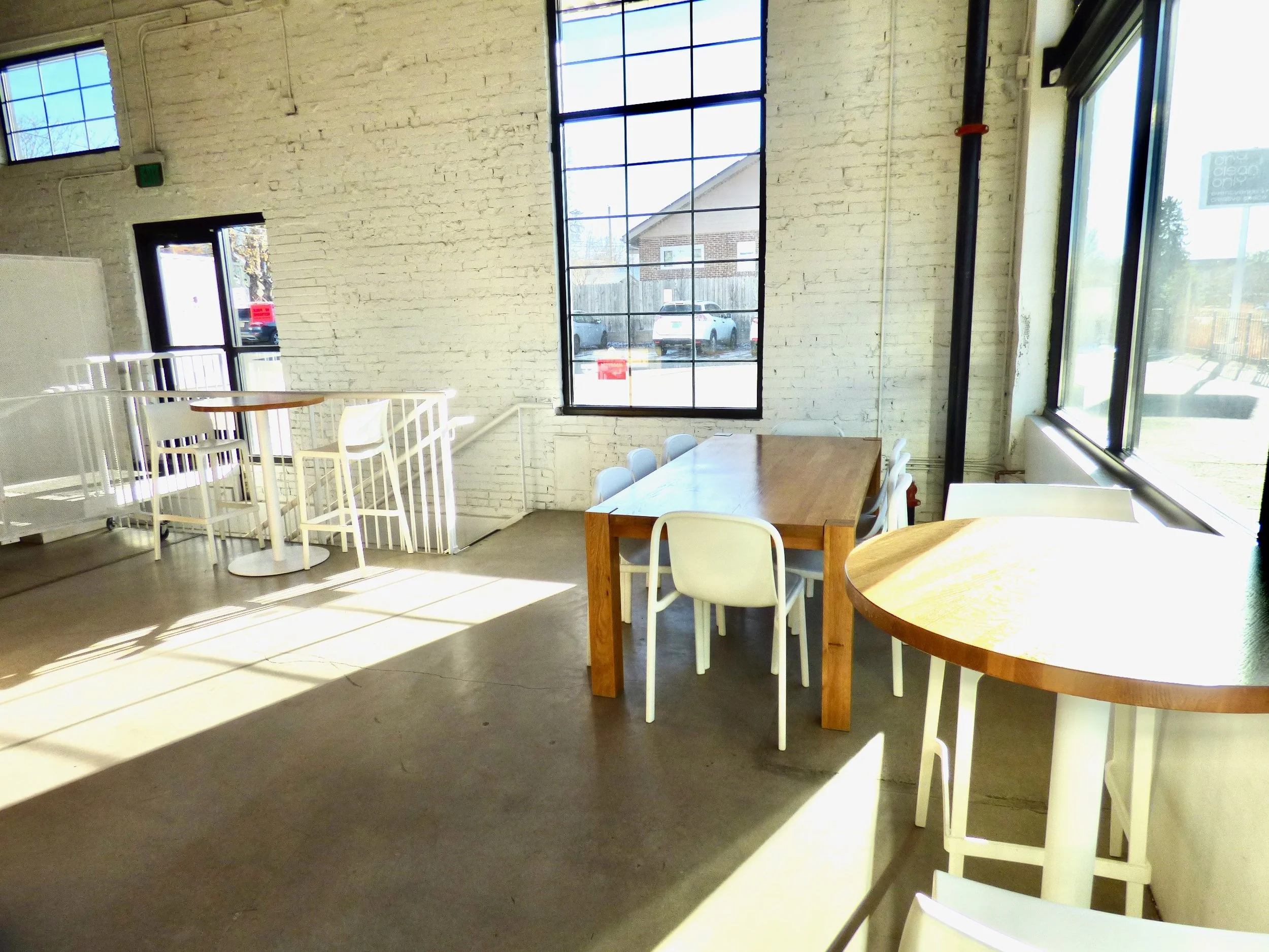 Bright interior of a cafe with white brick walls, large windows letting in sunlight, and various tables and chairs, including a wooden rectangular table with white chairs and high-top tables with white chairs at Hot Shot Coffee Shop, Denver, CO 80205