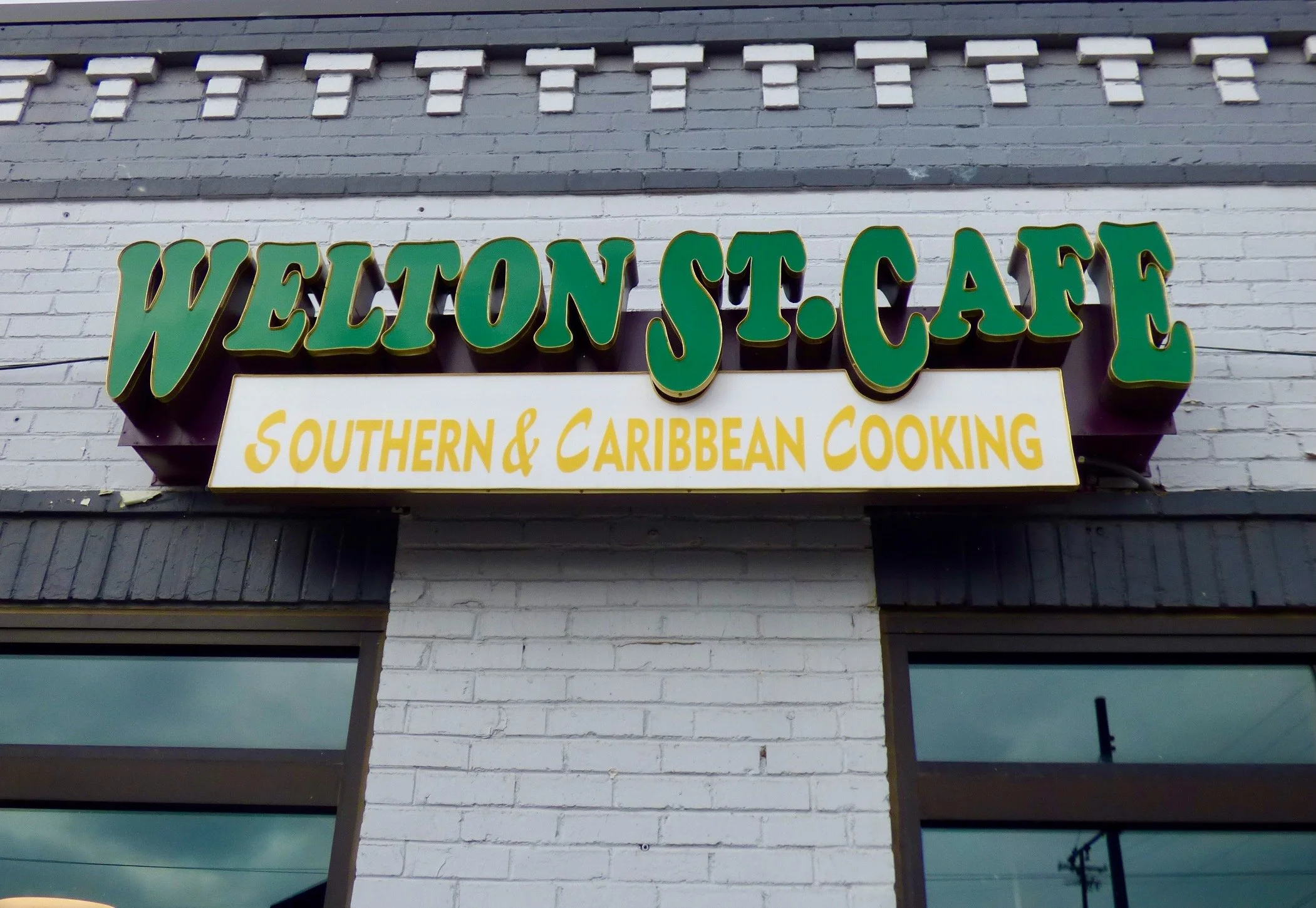 Sign for Welton St. Cafe with the subtitle Southern & Caribbean Cooking, mounted on a white brick wall above a window.