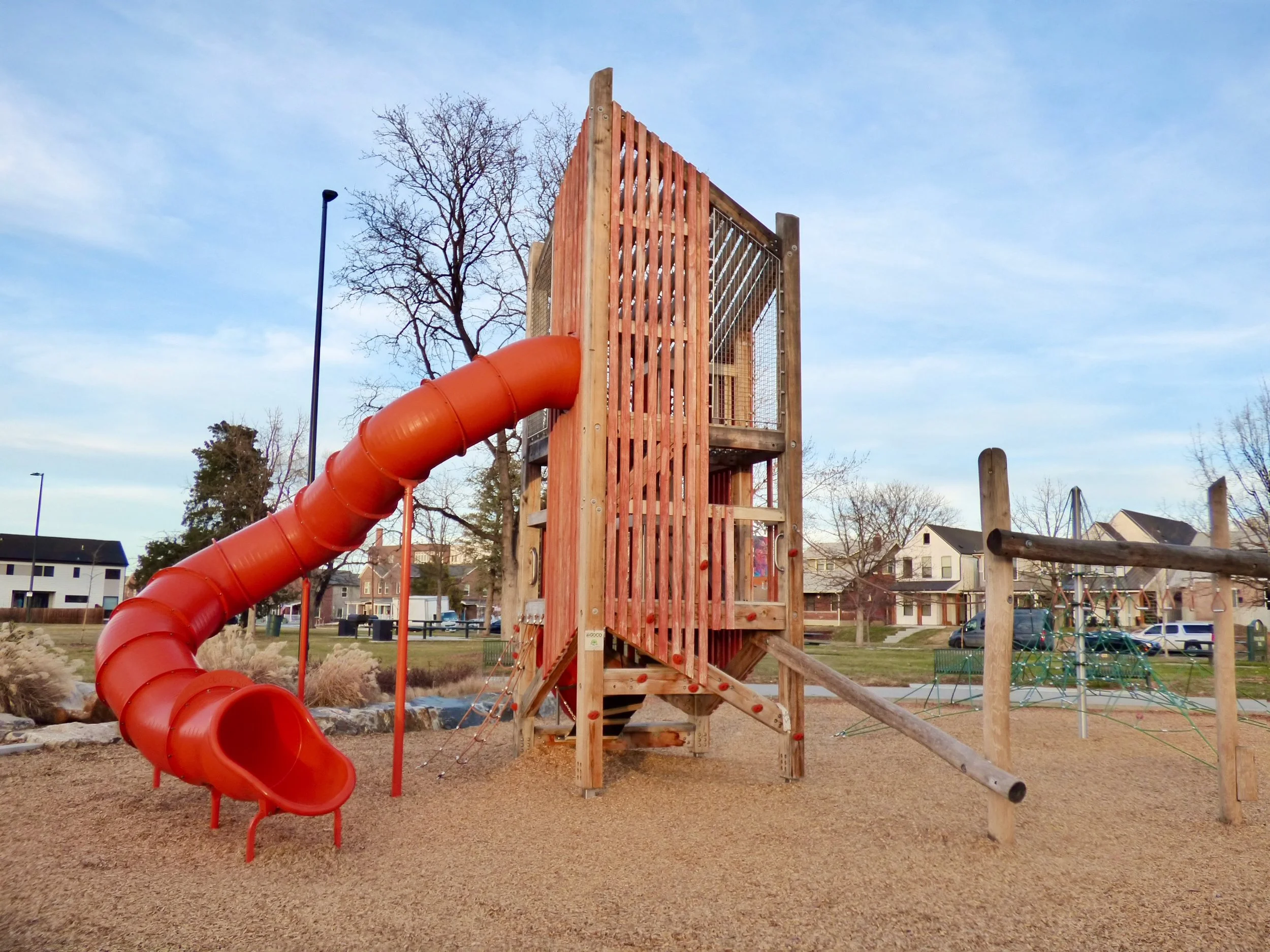 Wooden playground structure with a red spiral slide in a park with trees, houses, and a blue sky in the background in Russell Square Park, Denver, CO 80205.