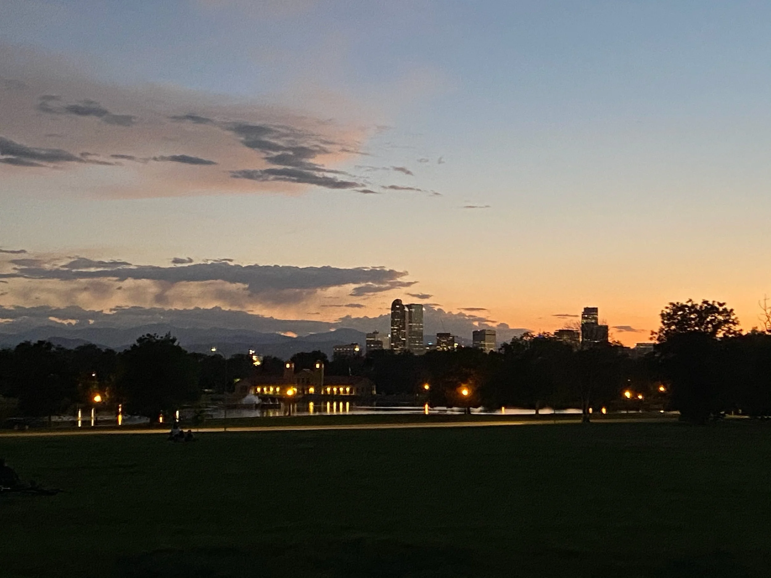 City skyline at dusk with high-rise buildings, some illuminated, visible behind trees in a park with water and a fountain in the foreground at City Park, Denver, CO.