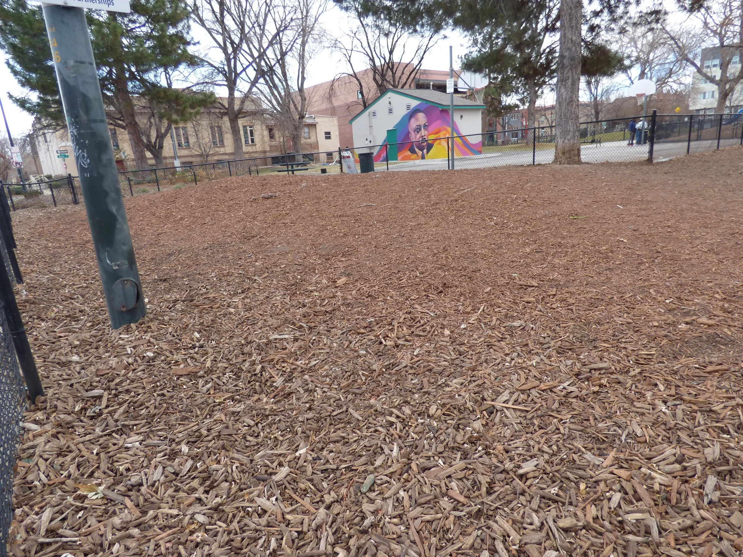 View of a playground with wood chip ground cover, a basketball hoop, a fenced court, trees, and buildings in the background. A colorful mural depicting a person's face is painted on the back of a small building.