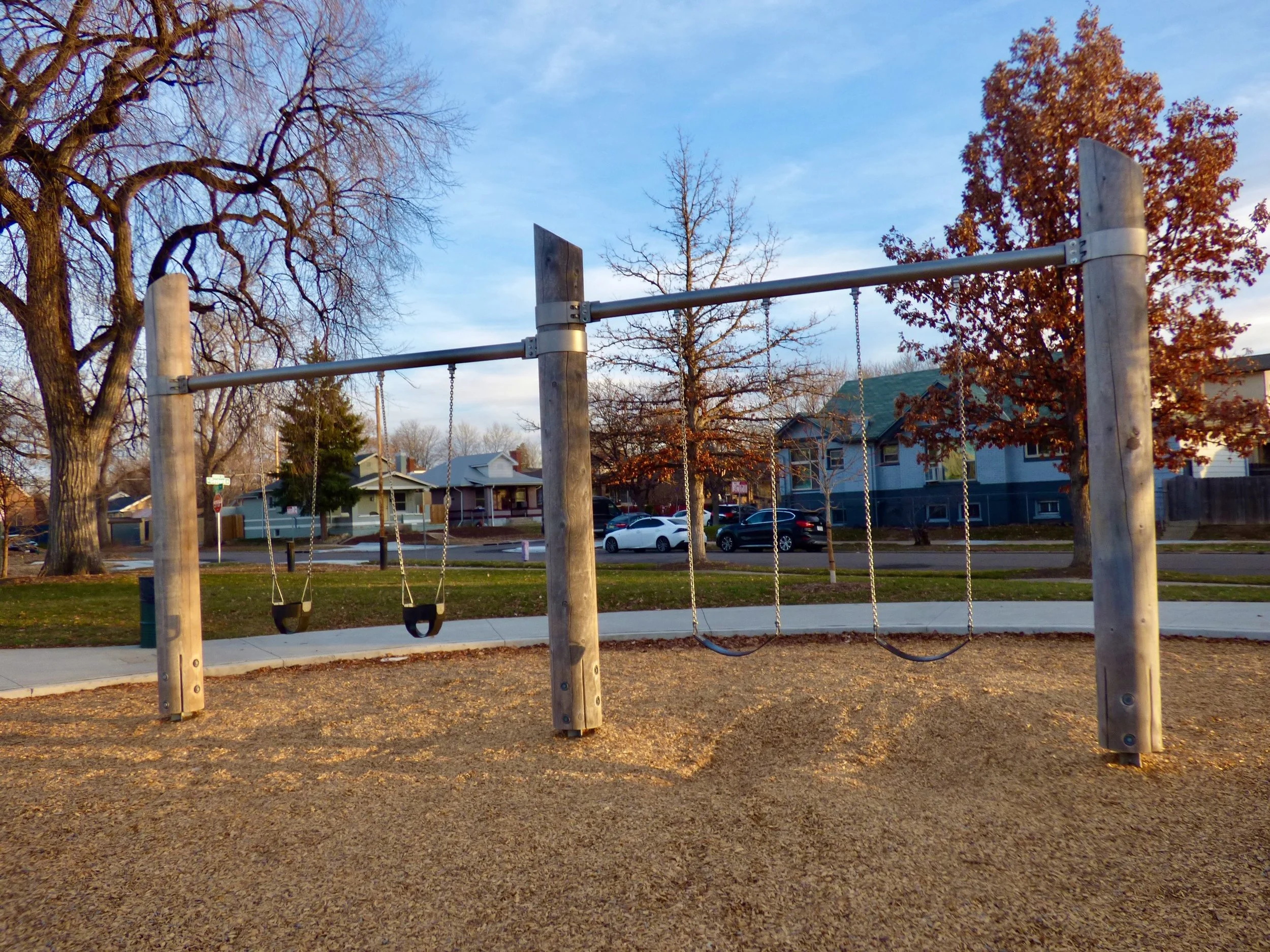 A playground swing set with three swings, two bucket swings on the left and two regular swings on the right, in a park with bare trees and residential houses in the background during late afternoon in Russell Square Park, Denver, CO 80205.