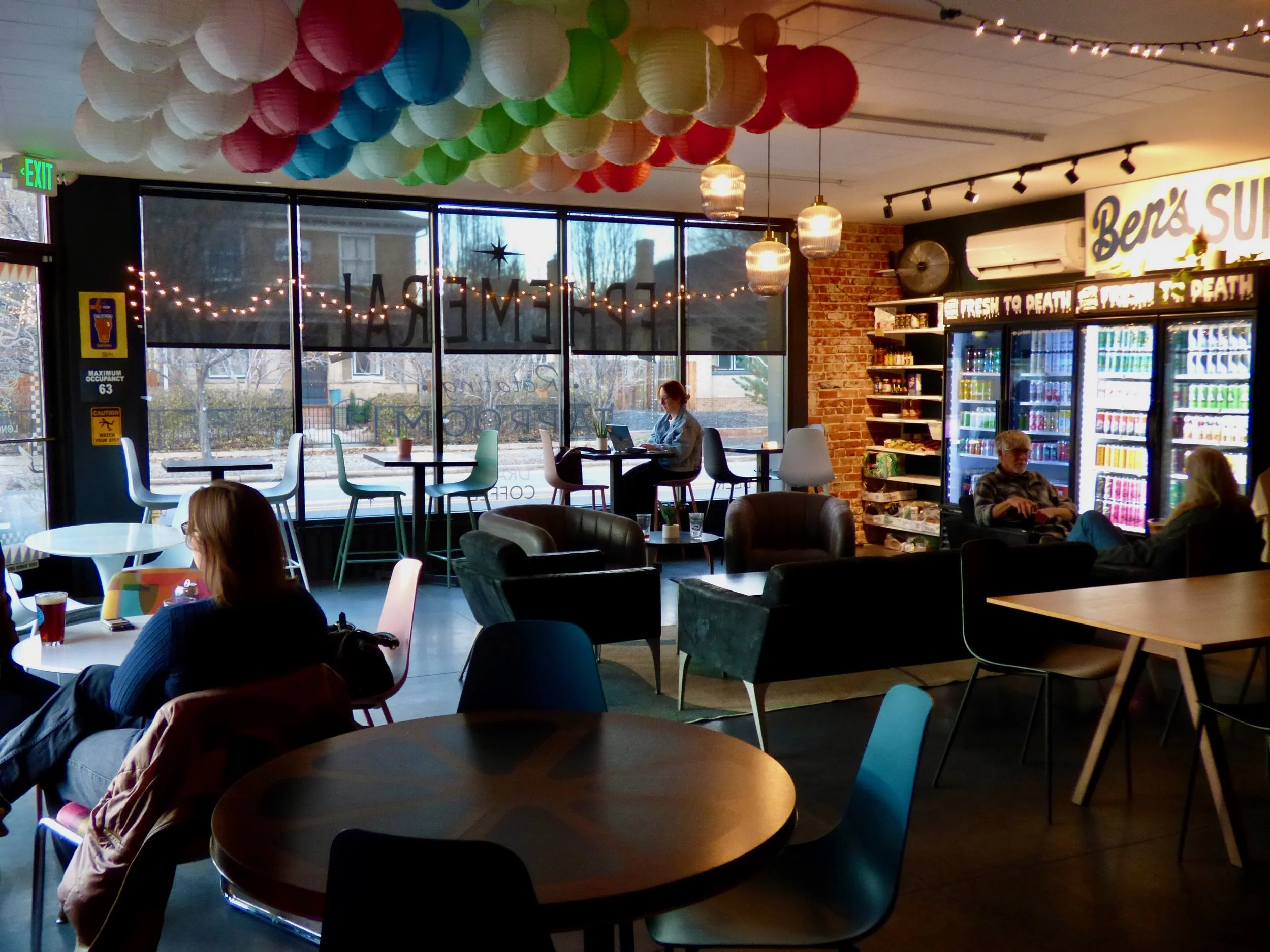 Interior of a coffee shop or café with colorful paper lantern decorations hanging from the ceiling, large windows with natural light, and customers seated at tables at Ephemeral Rotating Taproom, Denver, CO 80205.