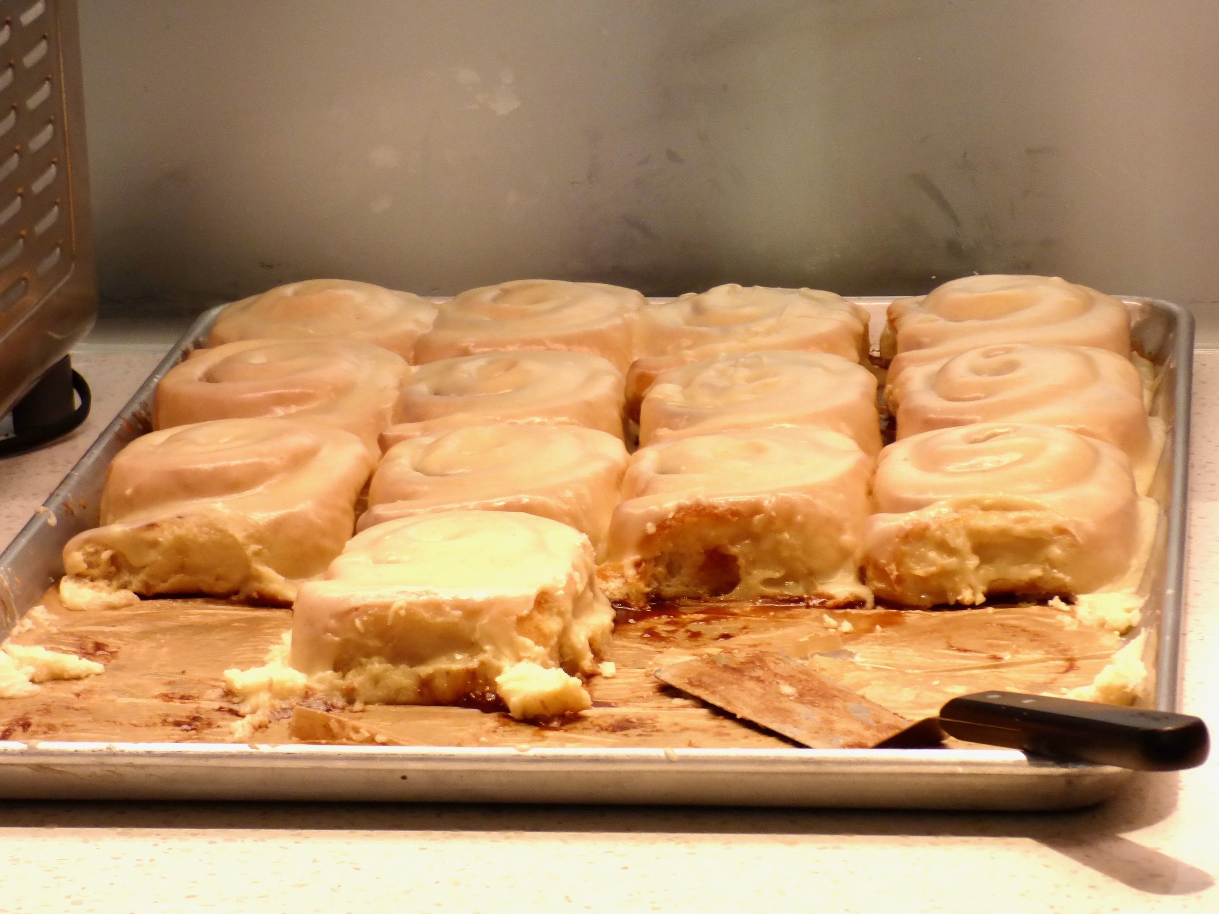 A metal tray filled with cinnamon rolls glazed with icing, some with a bite taken out, on a kitchen countertop at Elemental Bakery & Coffeehouse, Denver, CO 80205.