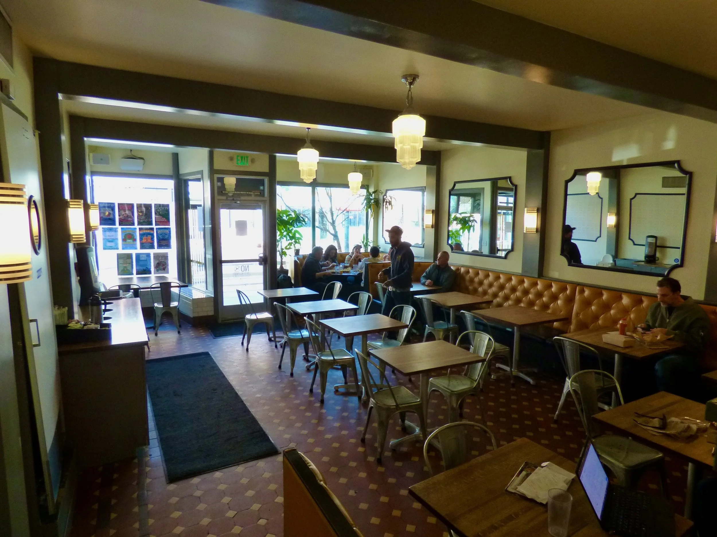 Interior of a cozy café or restaurant with wooden tables, metal chairs, a black doormat, mirrors on the wall, and customers seated while a server approaches.
