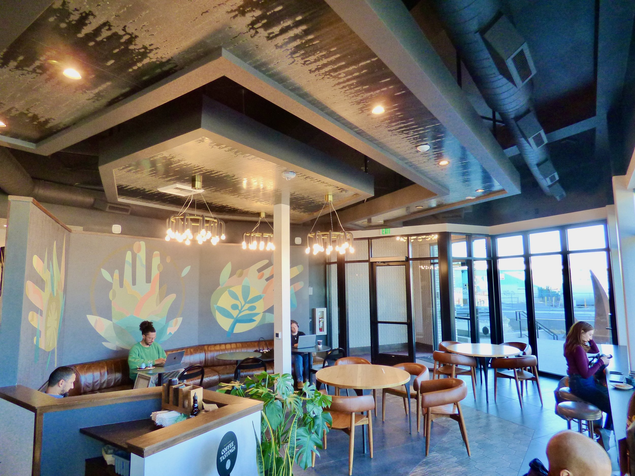 Interior of a modern coffee shop with large glass windows, wooden tables, leather seating, and hanging light fixtures, with three people using laptops and one person reading at Corvus Coffee Roasters, Denver, CO 80205.