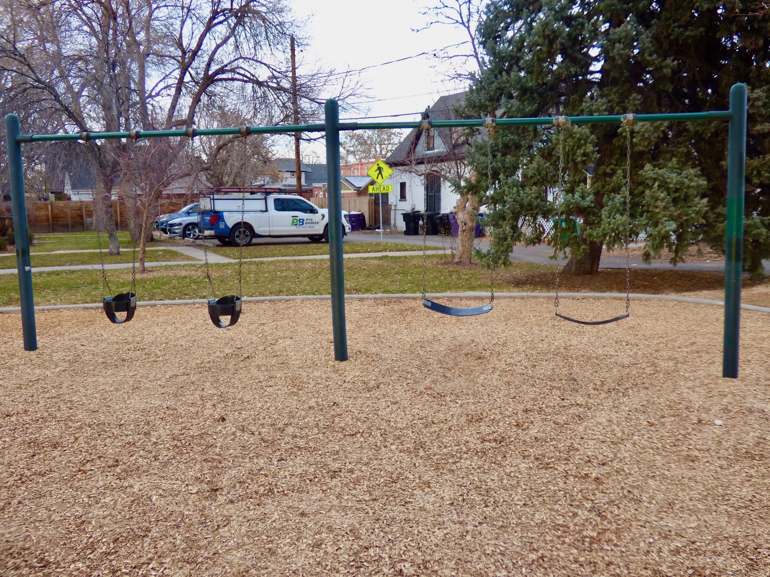 Empty swing set with four swings in a park, with trees, houses, and a parked city truck in the background at Frederick Douglas Park, Denver, CO 80205.