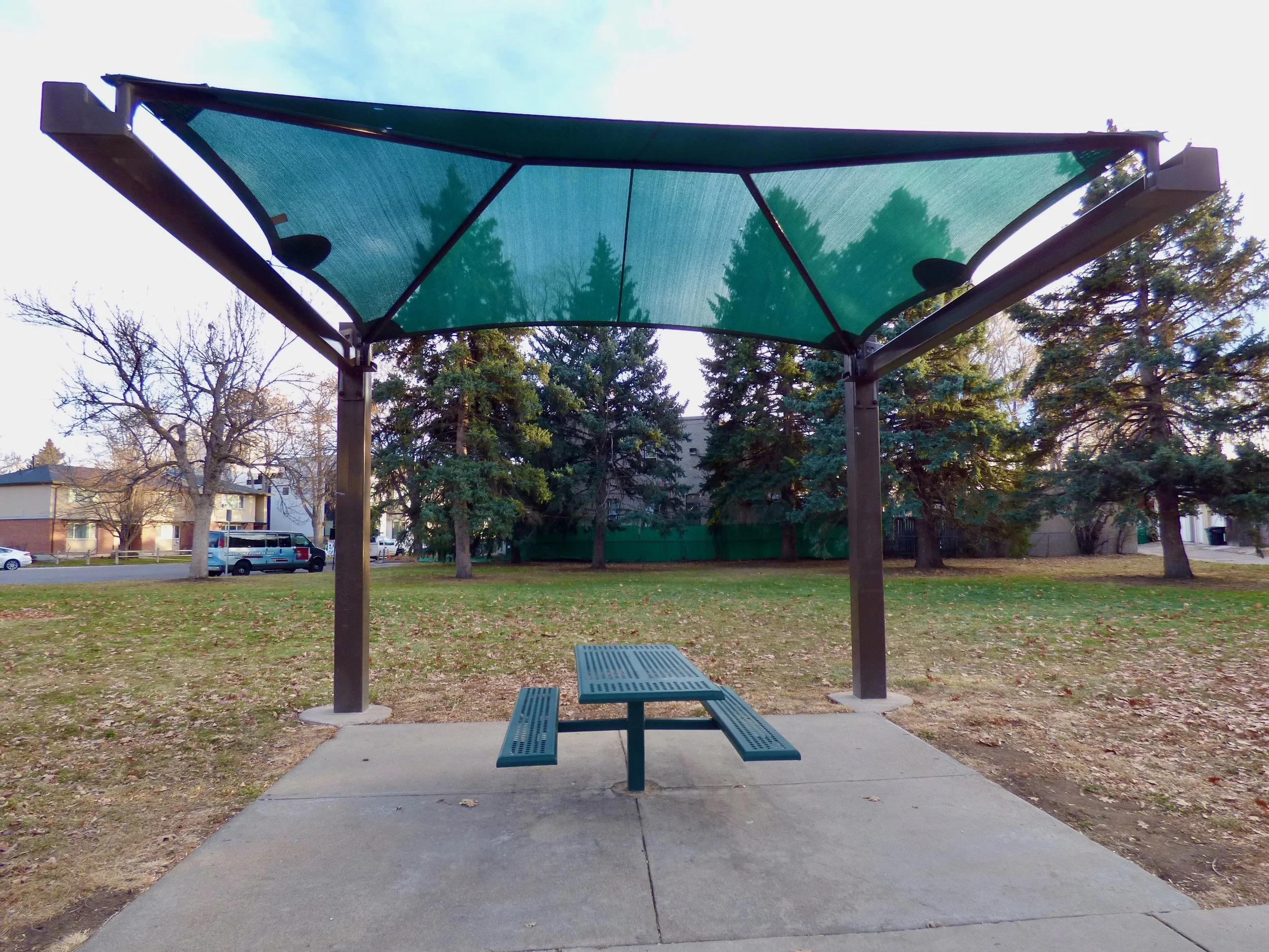 A park shelter with a green canopy shade structure and a picnic table on a concrete pad, surrounded by trees and grassy area at George Morrison Sr. Park, Denver, CO 80205.