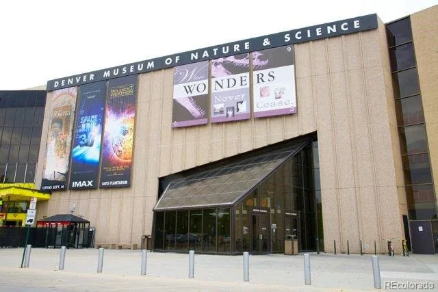 Exterior view of the Denver Museum of Nature & Science building with large banners advertising exhibits, a glass entrance, and a small parking area at Denver, CO.