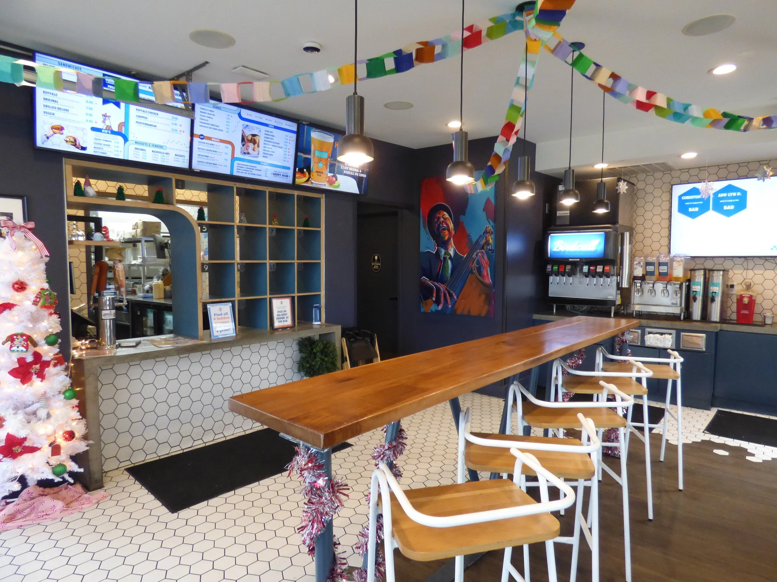 Interior of a modern casual restaurant decorated for Christmas, featuring a white Christmas tree, holiday garlands, a mural of a jazz musician, digital menu boards, a drink station, a wooden high-top table with chairs, and festive lighting.