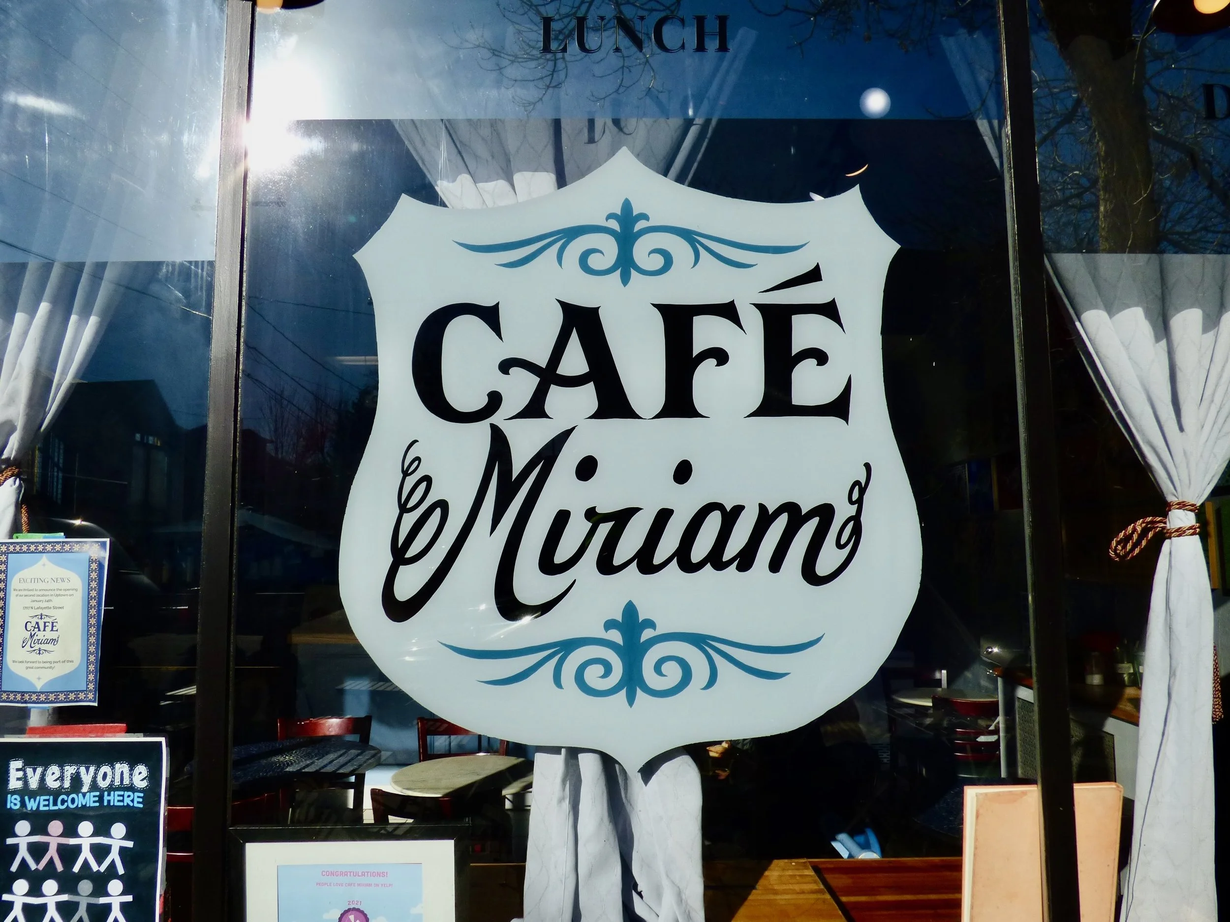 The front window of a cafe named Cafe Miuang with a large decorative sign in the center, reflecting sunlight and showing a dining area inside at Cafe Miriam, Denver, CO 80205.