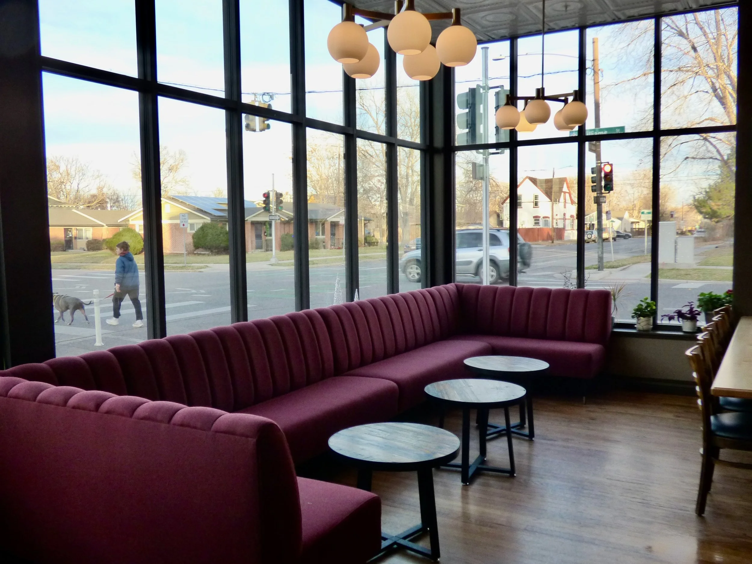 Interior of a cafe with a large pink velvet sectional sofa near a window, with three small round tables in front at Whit's End, Denver, CO 80205.