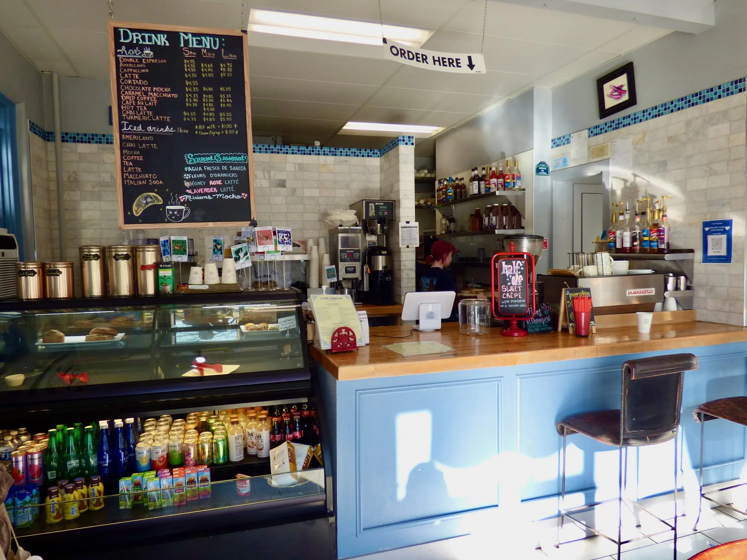 Inside a cozy cafe with a counter, a menu board, a glass display of drinks and snacks, and a barista preparing beverages. The cafe has a white-tiled wall, a light blue counter, and seating with bar stools at Weathervane Cafe, Denver, CO.