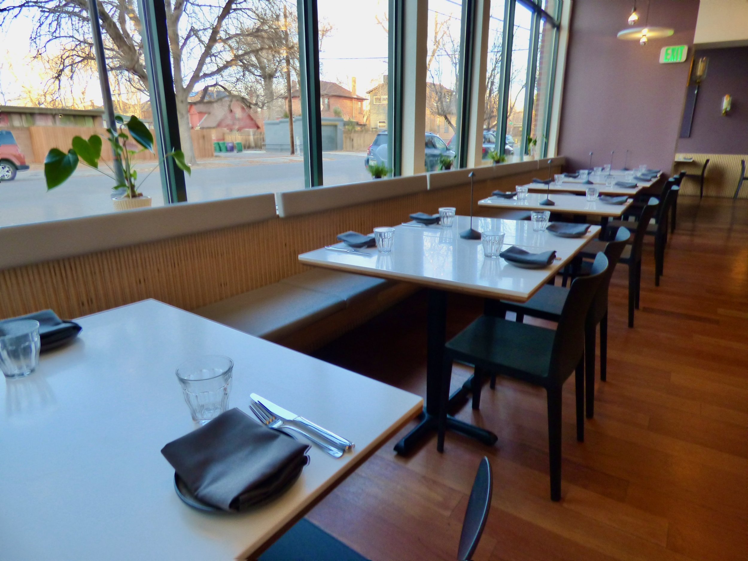 Empty restaurant table arranged with black napkins, silverware, and glasses, set along a large window with outdoor view, inside a modern dining space with wood flooring and subdued lighting at Point Easy, Denver, CO 80205.