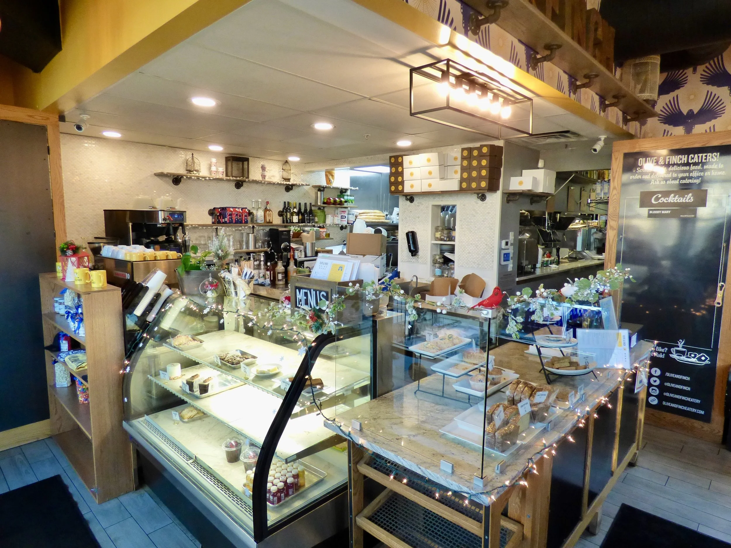 Interior of a cozy cafe with a display case filled with desserts, a counter with coffee machines and bottles, and a festive decor with string lights and holiday adornments at Olive & Finch, Denver, CO.