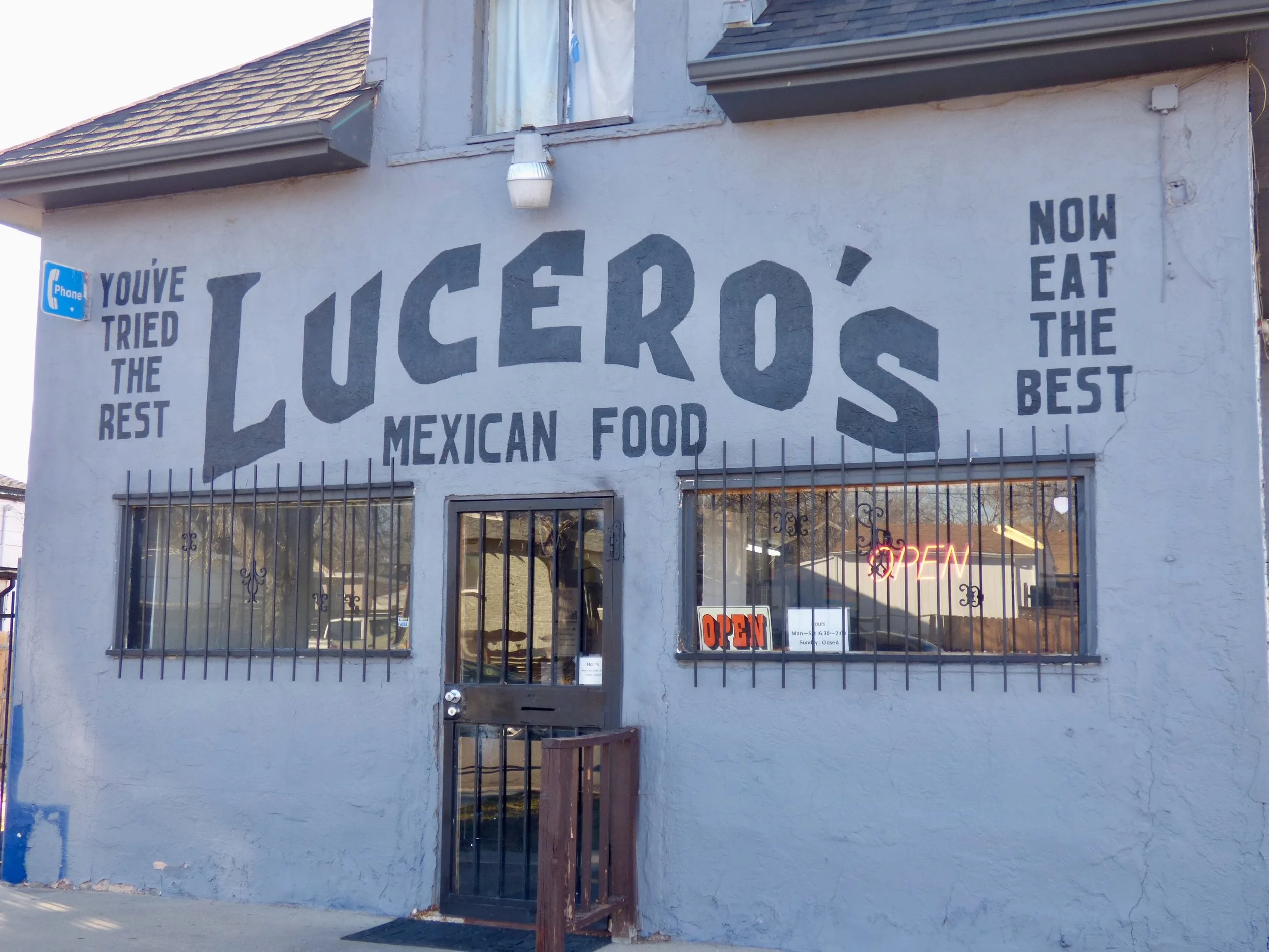Exterior of a Mexican food restaurant with a large painted sign reading 'LUCERO'S MEXICAN FOOD' and slogans 'YOU'VE TRIED THE REST' and 'NOW EAT THE BEST'. The building has barred windows and a neon 'OPEN' sign in the window.