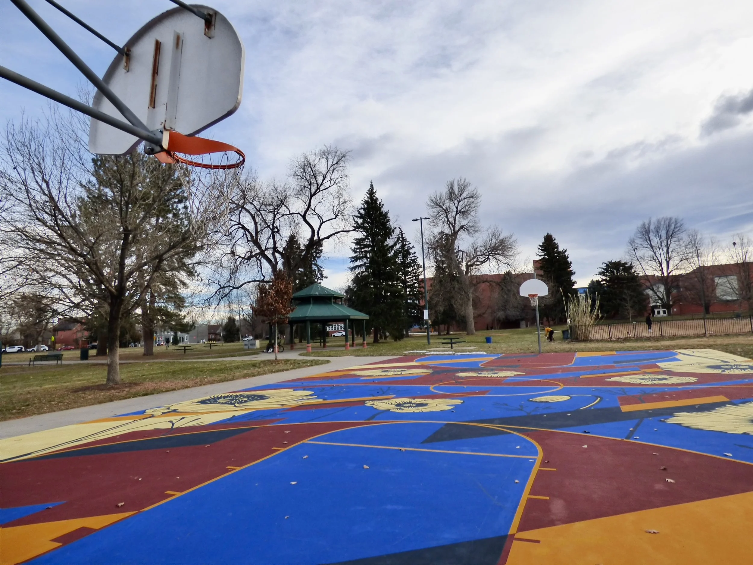 Empty outdoor basketball court in a park with colorful artwork on the ground, surrounded by trees, a pavilion, and a fence under a cloudy sky at Fuller Park, Denver, CO 80205.