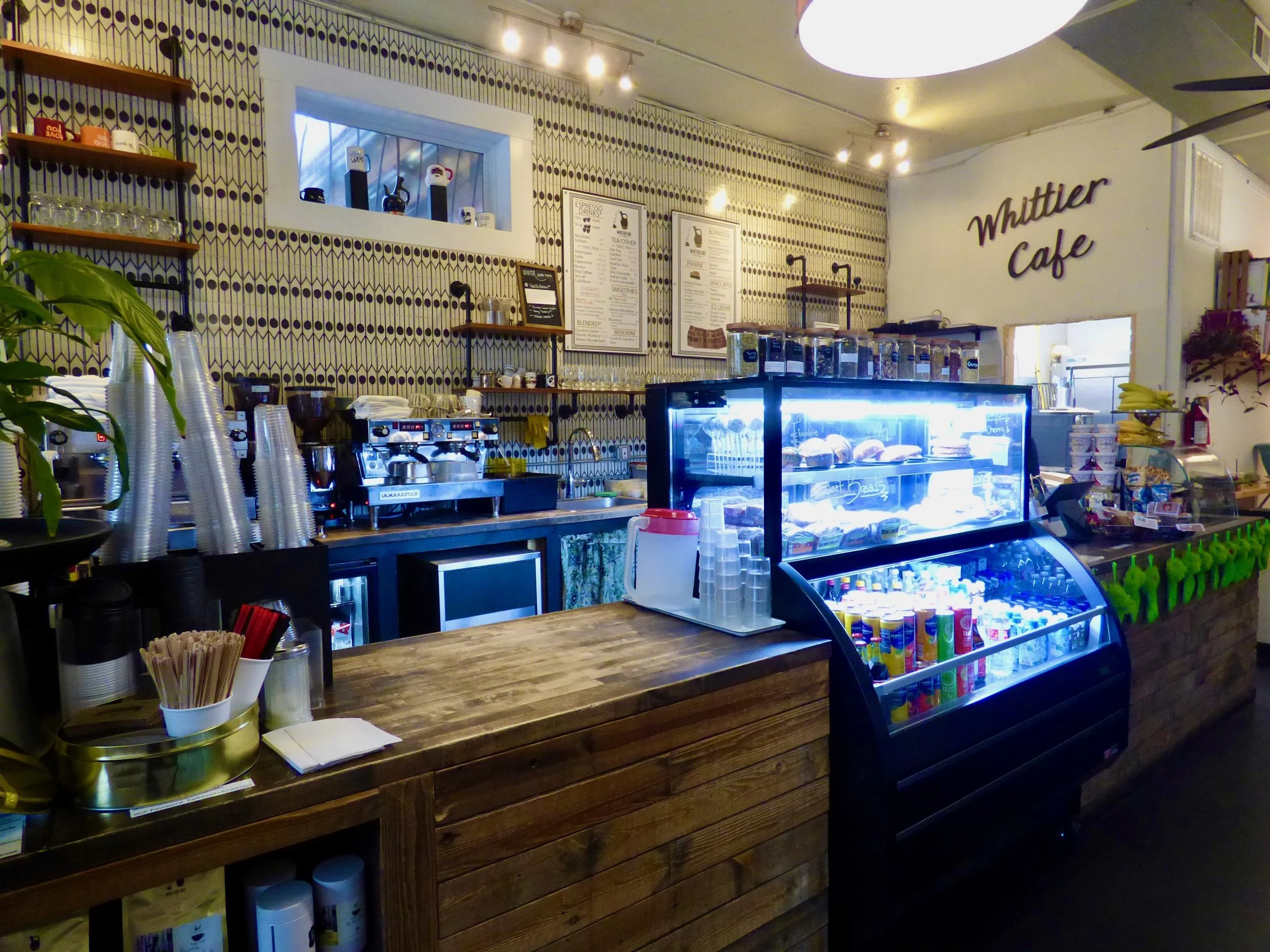 Interior of Whittler Cafe with a wooden counter, shelves with cups, a coffee machine, and a refrigerated display case with drinks and desserts at Cafe Miriam, Denver, CO 80205.