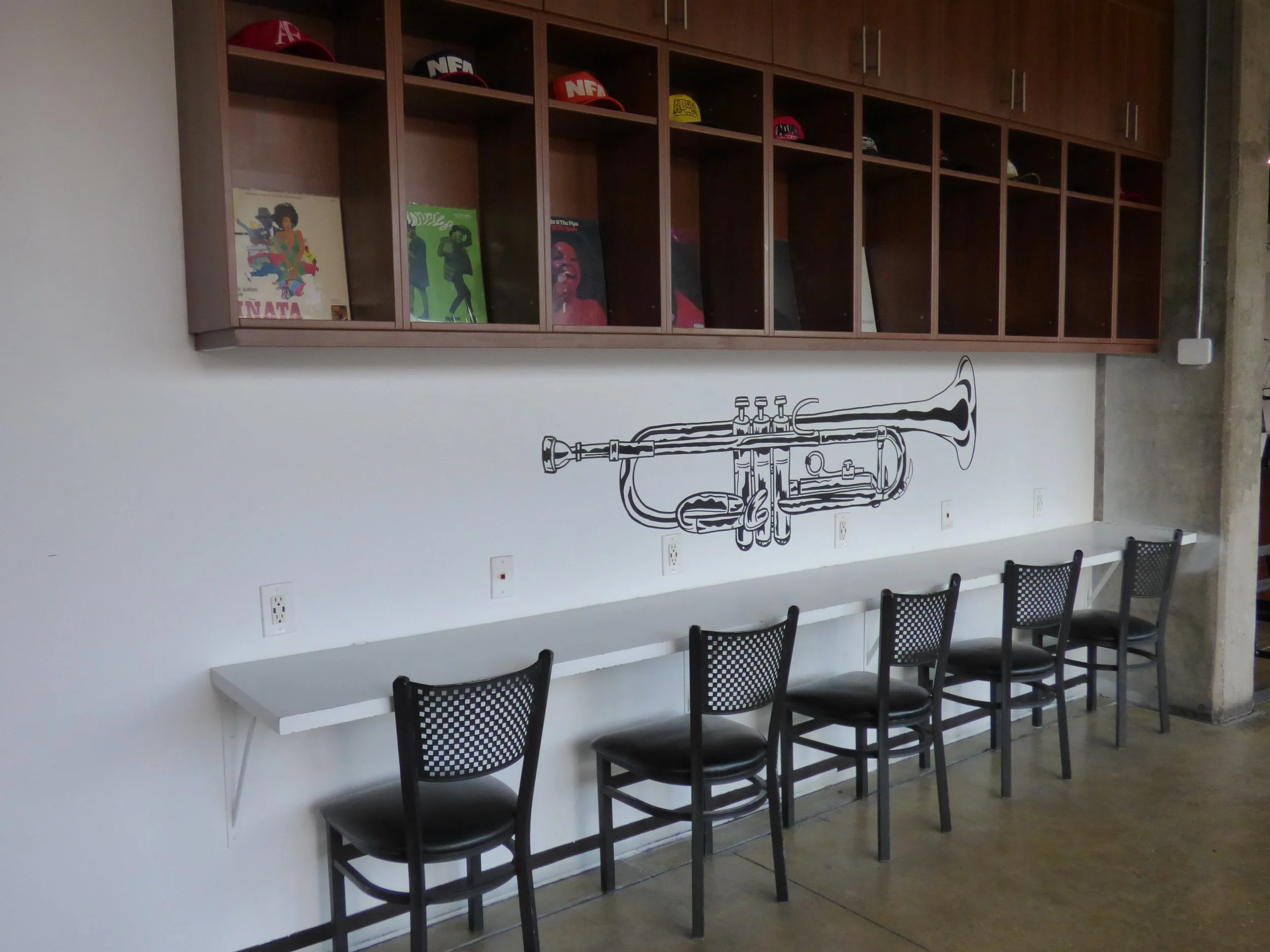 Bar wall with a black and white trumpet mural, wooden shelves with hats and vinyl records, and four black chairs with a white counter Fifth Coffee House, Denver, CO 80205.