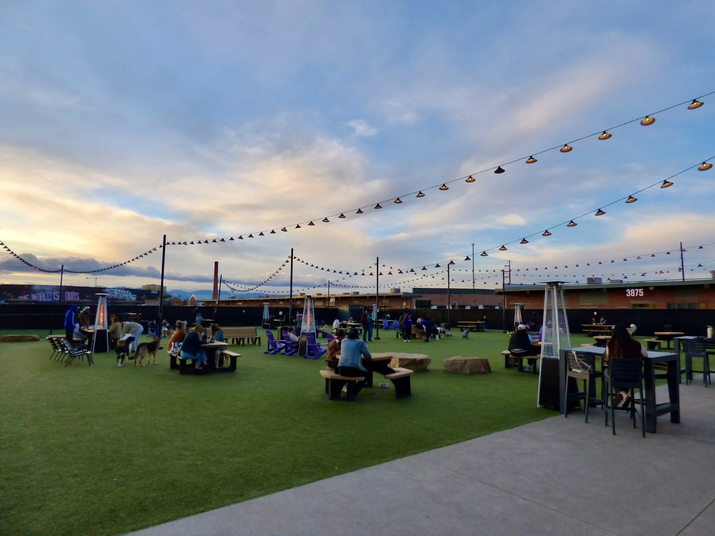 An outdoor rooftop gathering area at dusk with string lights, outdoor heaters, tables, and people socializing on a green artificial grass surface at Skiptown, Denver, CO 80205.
