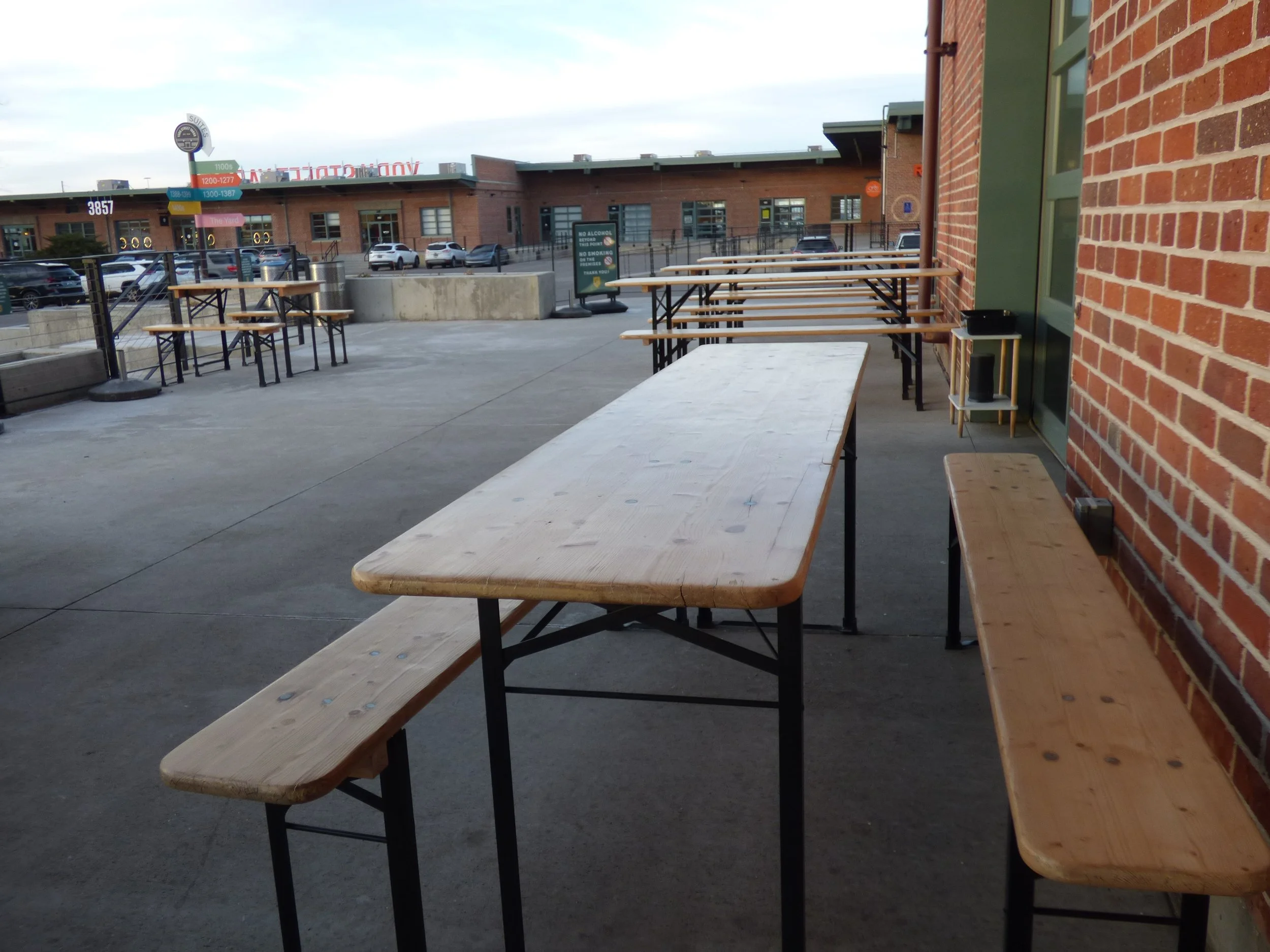 Empty outdoor patio area with wooden tables and benches, brick wall on the right, parking lot and buildings in the background at Cohesion Brewery, Denver, CO 80205.
