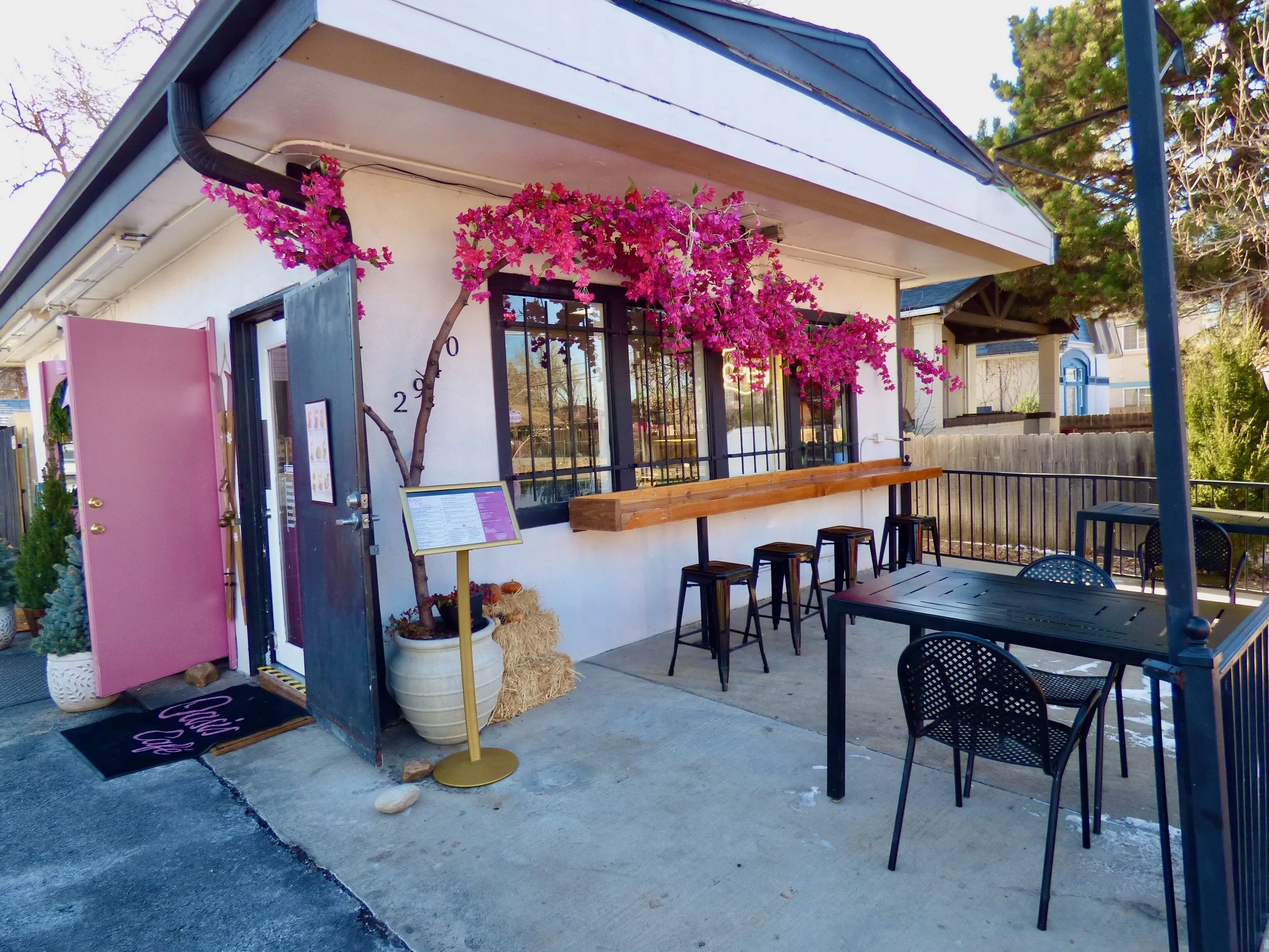 Outdoor seating area of a cafe with pink bougainvillea flowers and pink door, black tables and chairs, and a wooden bar counter at Oasis Cafe & Grill, Denver, CO 80205.