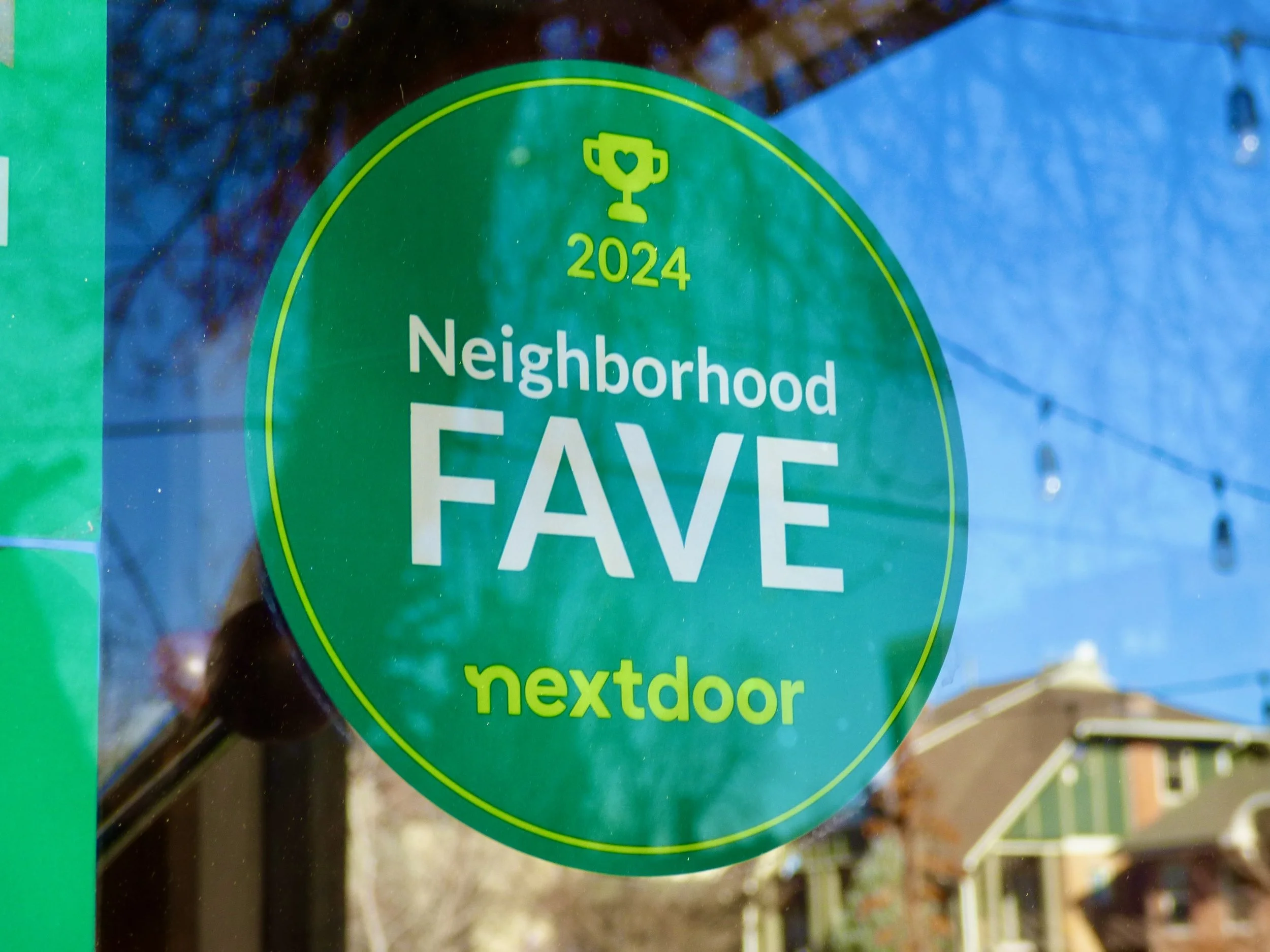 Green sticker on glass window reads 'Neighborhood FAVE nextdoor 2024' with a trophy icon above, and a house with trees and power lines are visible in the background at The Plimoth, Denver, CO 80205.